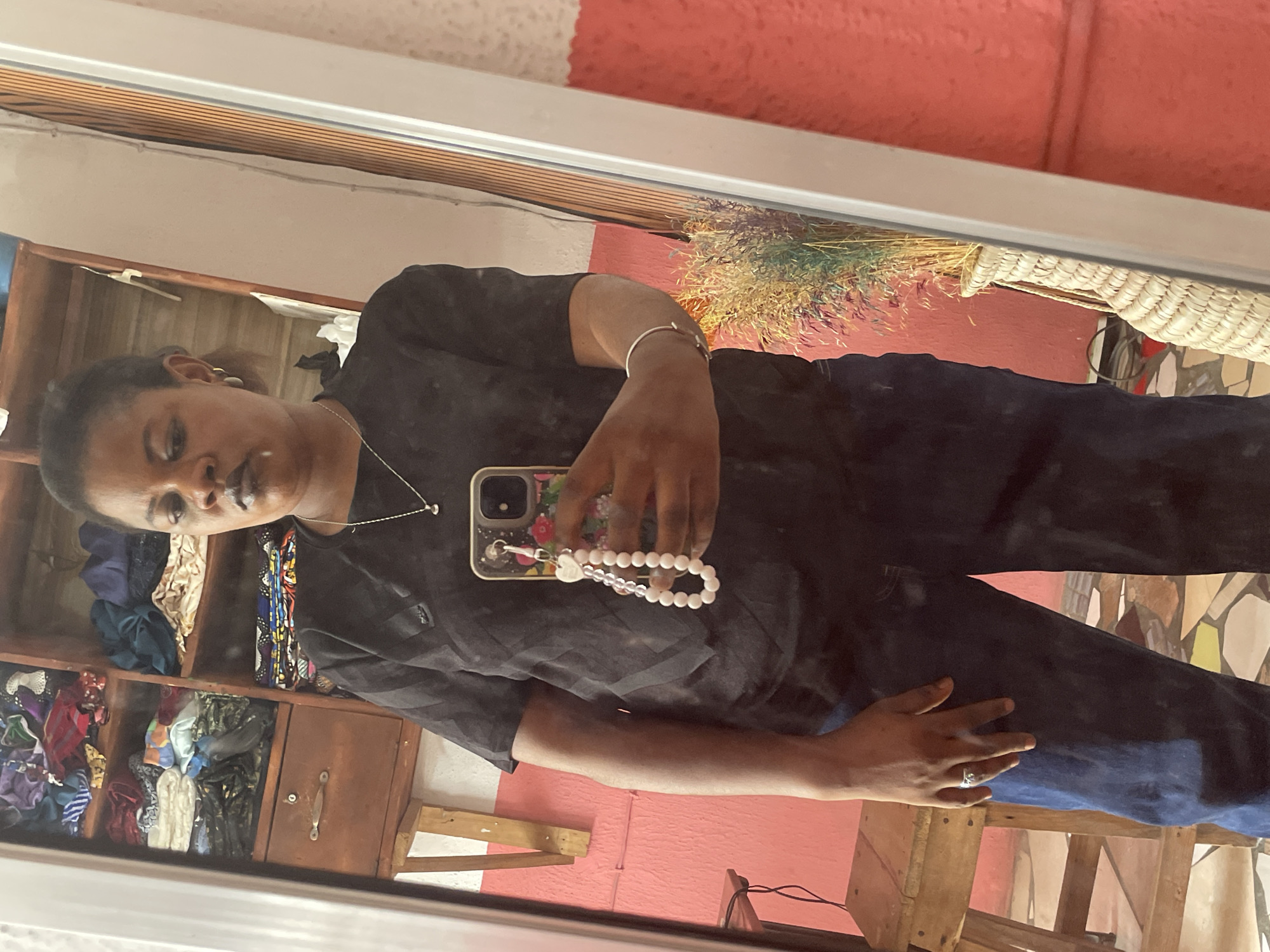 A woman is reflected in a mirror, holding a smartphone with a beaded charm, wearing a silver bracelet and necklace. She has on a black shirt and blue jeans. Behind her is a shelf filled with textiles. Adjacent to her is a basket of dried plants. She appears to be indoors, the scene possibly within a shop. The location appears to be in Badagry, Nigeria.