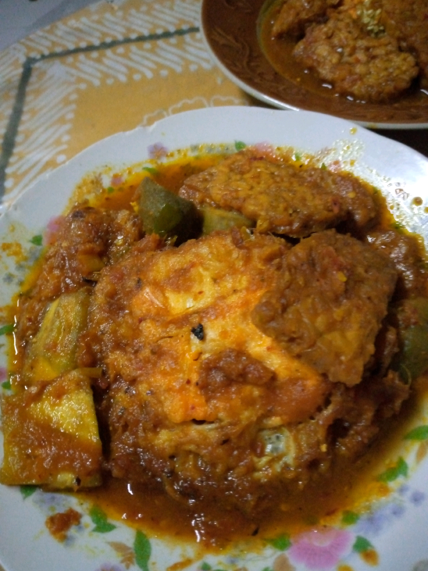In Sumberwono, Indonesia, a close-up shot captures a plate of food, possibly an Indonesian dish like *telur balado* or a similar curry with fried eggs and vegetables. The food is presented in a thick, reddish-brown sauce, with visible pieces of fried egg, some green vegetables, and possibly onion. The plate is white with a floral pattern along the rim. In the background, a glimpse of a table with a yellow and white patterned cloth is visible, and another dish with similar food is out of focus.