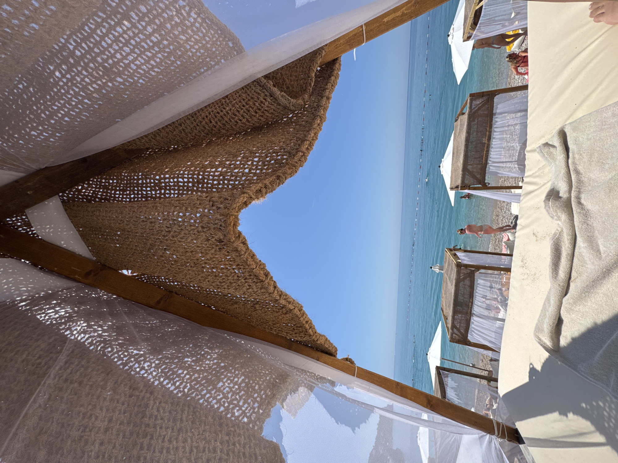 The image depicts a bright, sunny day at a beach in Vrba, Montenegro, viewed from within a beach cabana. In the immediate foreground, a light-colored, possibly beige, cushioned sunbed or daybed occupies the lower portion of the frame, with a matching towel draped over it and the edge of a foot visible in the bottom right corner. Above, the cabana's structure features rustic wooden beams supporting a roof composed of two layers: an inner sheer white mesh fabric and an outer coarse, woven brown material with a netting pattern.
Beyond the cabana, a pebble beach extends towards the water. Scattered along the shore are several other similar cabanas with white fabric canopies and white beach umbrellas providing shade. People are present on the beach and in the clear, calm, turquoise-to-deep-blue sea. On the right side of the beach, a woman in a light-colored bikini stands facing away from the viewer, near the water's edge. Another person in a red swimsuit is partially visible to her left, closer to the water. Further out in the water, a line of buoys marks a swimming area, with additional individuals swimming or wading. The horizon is clear, where the blue sea meets an expanse of bright, cloudless blue sky.