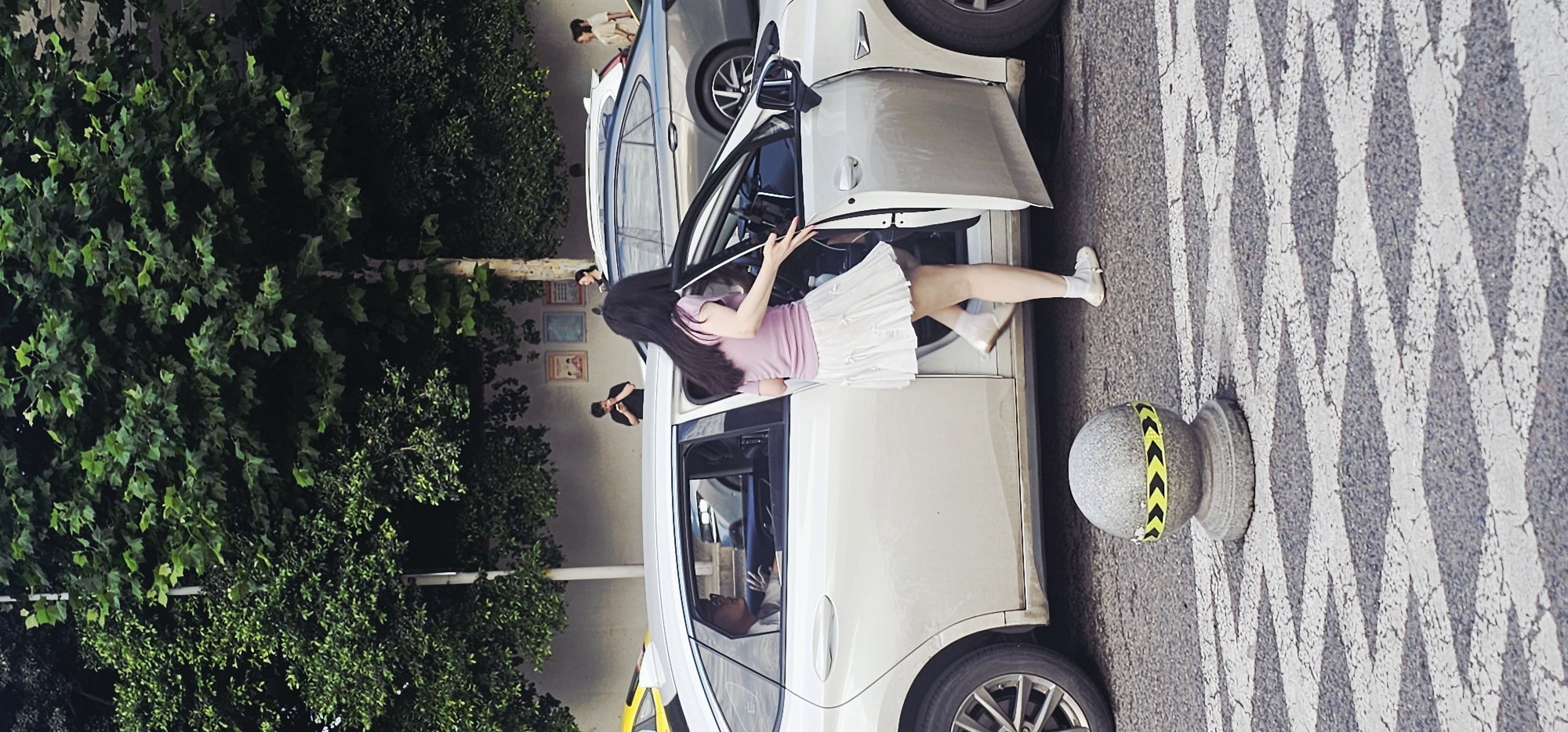 A daytime shot shows a woman exiting a white car on a city street in Yuzhong District, China. The woman has long dark hair, and wears a pink shirt, white skirt, and white shoes. She is stepping out of the driver's side of the car. A grey concrete ball with yellow and black chevrons is on the ground next to the car. Behind the car is another car, and people are visible in the background near a wall with pictures. Lush green trees are growing along the left side of the frame. The street has zebra striping.