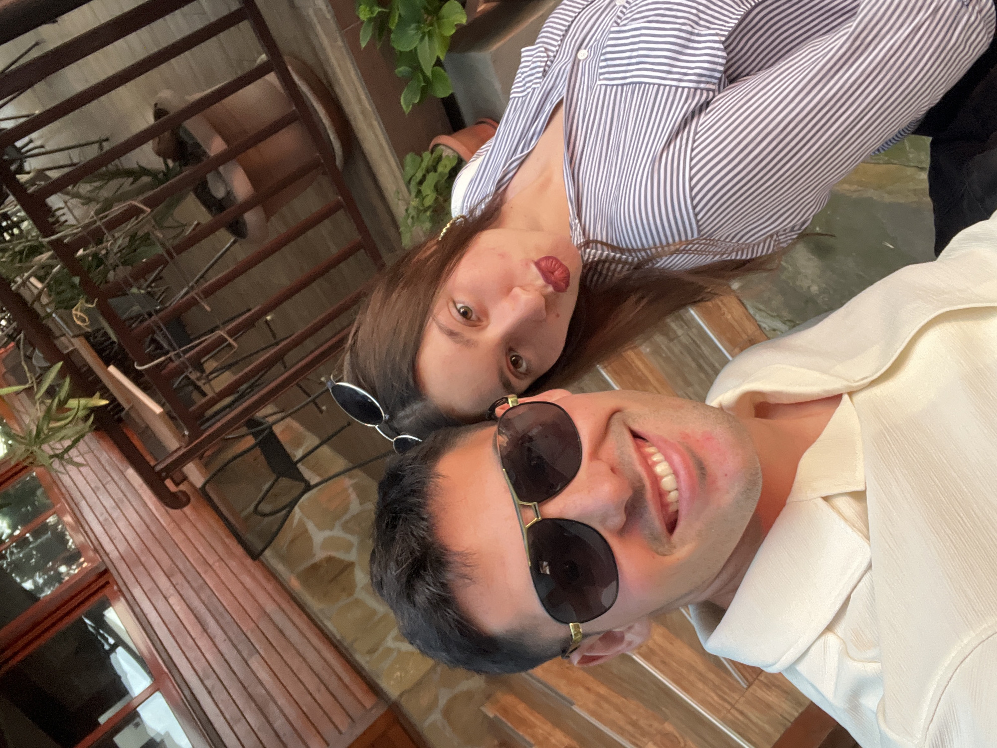 A tilted, close-up shot features two individuals, a man and a woman, seemingly posing for a selfie outdoors. The man, in the foreground on the right, smiles broadly, revealing his teeth. He has short dark hair, a light stubble, and wears aviator-style sunglasses with gold frames, along with a light-colored, possibly cream or off-white, collared shirt.
Behind and to the left of the man is the woman. She has long brown hair, dark eyes, and is wearing a striped blue and white button-down shirt. She wears round, dark-lensed sunglasses perched on her head and makes a playful face, with her tongue slightly extended.
The background suggests an outdoor setting, possibly a patio or balcony. To the left, there's dark wooden fencing or railings and green plants. Further back, visible through the railings, are more plants in pots and wooden siding on a building. The floor surface under the man appears to be light-colored tiles or stone. The lighting indicates it's daytime. No visible text is present in the image.
