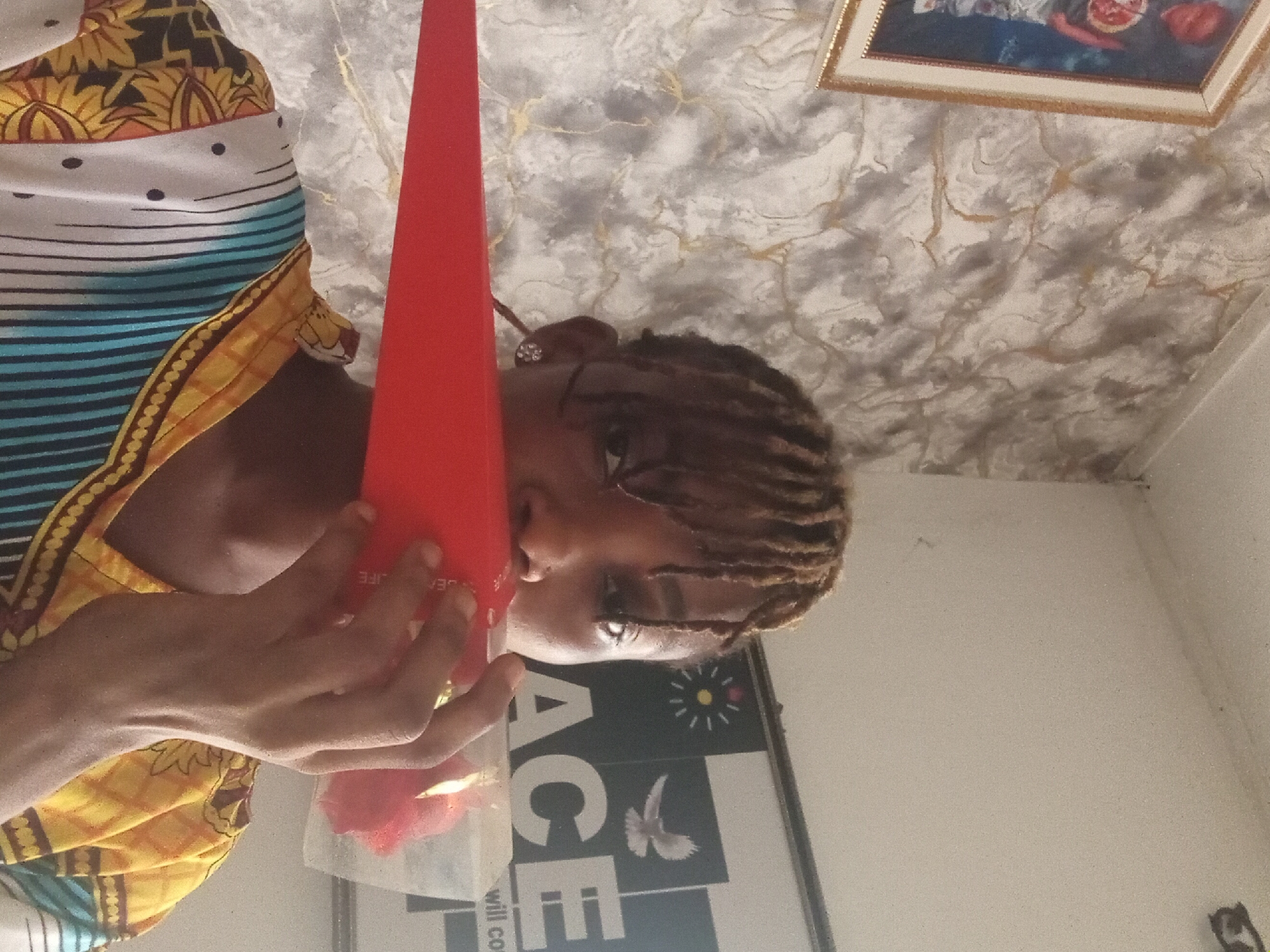 The image depicts a person with braided hair, adorned with an earring, holding a red, conical object. The person is wearing a patterned shirt with a mix of yellow, white, and blue stripes. In the background, there is a poster with the word "ACE" and a picture of a dove, along with a framed picture on the ceiling. The location is Unknown, Unknown.