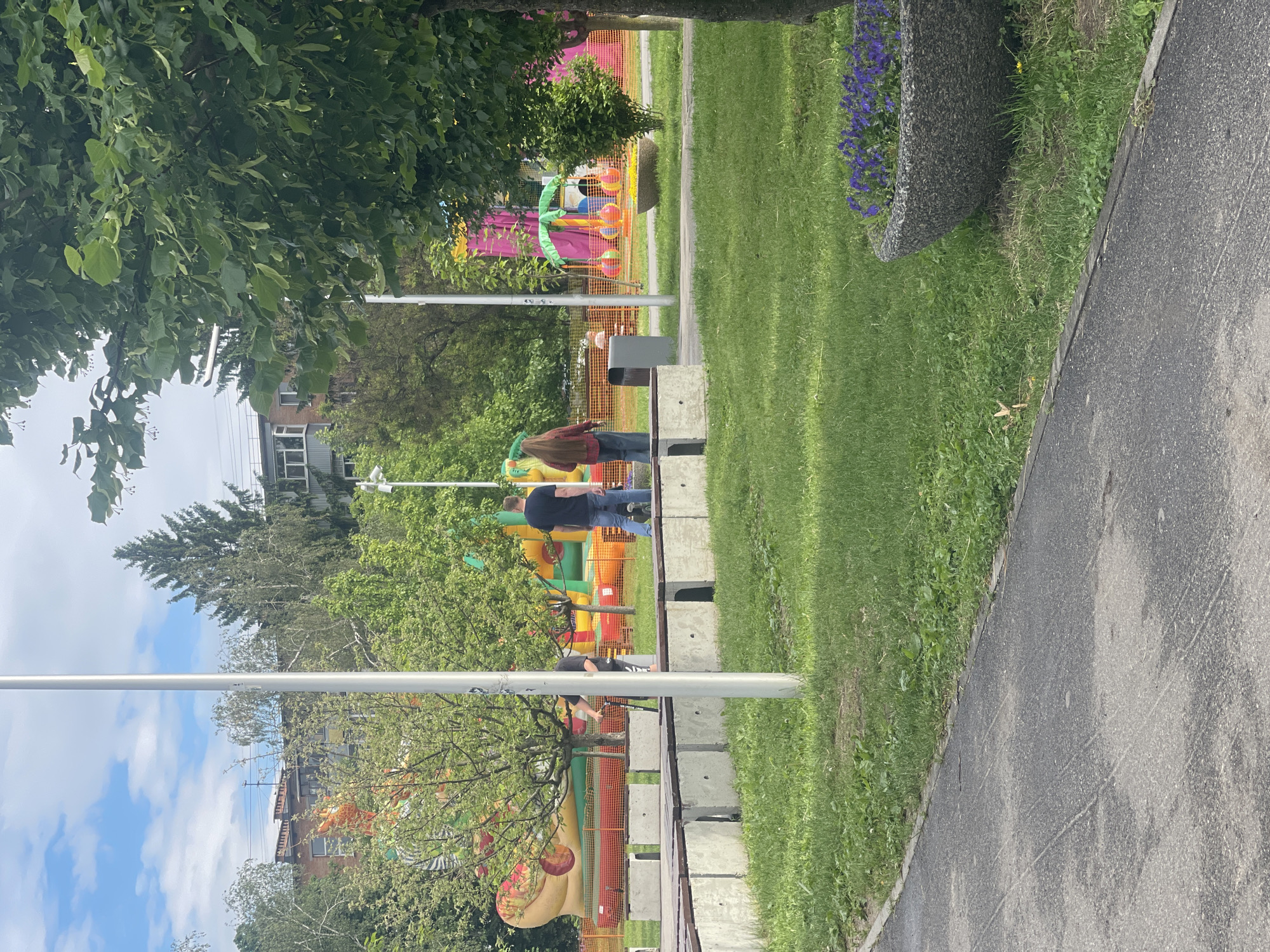 This image, taken in Poltava, Ukraine, depicts an outdoor recreational area, likely a park, under a partly cloudy sky. The scene is characterized by lush greenery and a vibrant children's play zone.
In the foreground, a dark asphalt path curves along the right side of the frame, bordered by a strip of green grass. To the left of the path, a series of low, grey concrete blocks or benches form a barrier. Large green trees with dense foliage obscure parts of the upper left.
In the mid-ground, several individuals are present near the concrete barrier. Two adults, seen from behind, are standing; one wears a dark blue top and jeans, while the other is dressed in a reddish-brown plaid shirt and jeans. Further along the barrier, another person in a black t-shirt is bending down.
Beyond the concrete barrier and some orange mesh safety fencing, the area transforms into a colorful children's play zone. It features multiple inflatable structures, including a multi-colored bouncy castle (red, yellow, green, blue), a large yellow and orange inflatable, a green inflatable palm tree, and large, colorful inflatable balls. An inflatable animal figure, resembling a crocodile, is also visible. These elements suggest an active children's event, a fair, or a dedicated play area.
The background is filled with numerous green trees, through which glimpses of multi-story buildings can be seen, indicating an urban or suburban environment. A large, textured grey planter filled with purple flowers is situated in the upper right. Tall, slender white poles are also scattered throughout the scene. The natural light indicates it is daytime, likely morning or afternoon, with a generally calm and leisurely atmosphere. No discernible text is visible within the image.