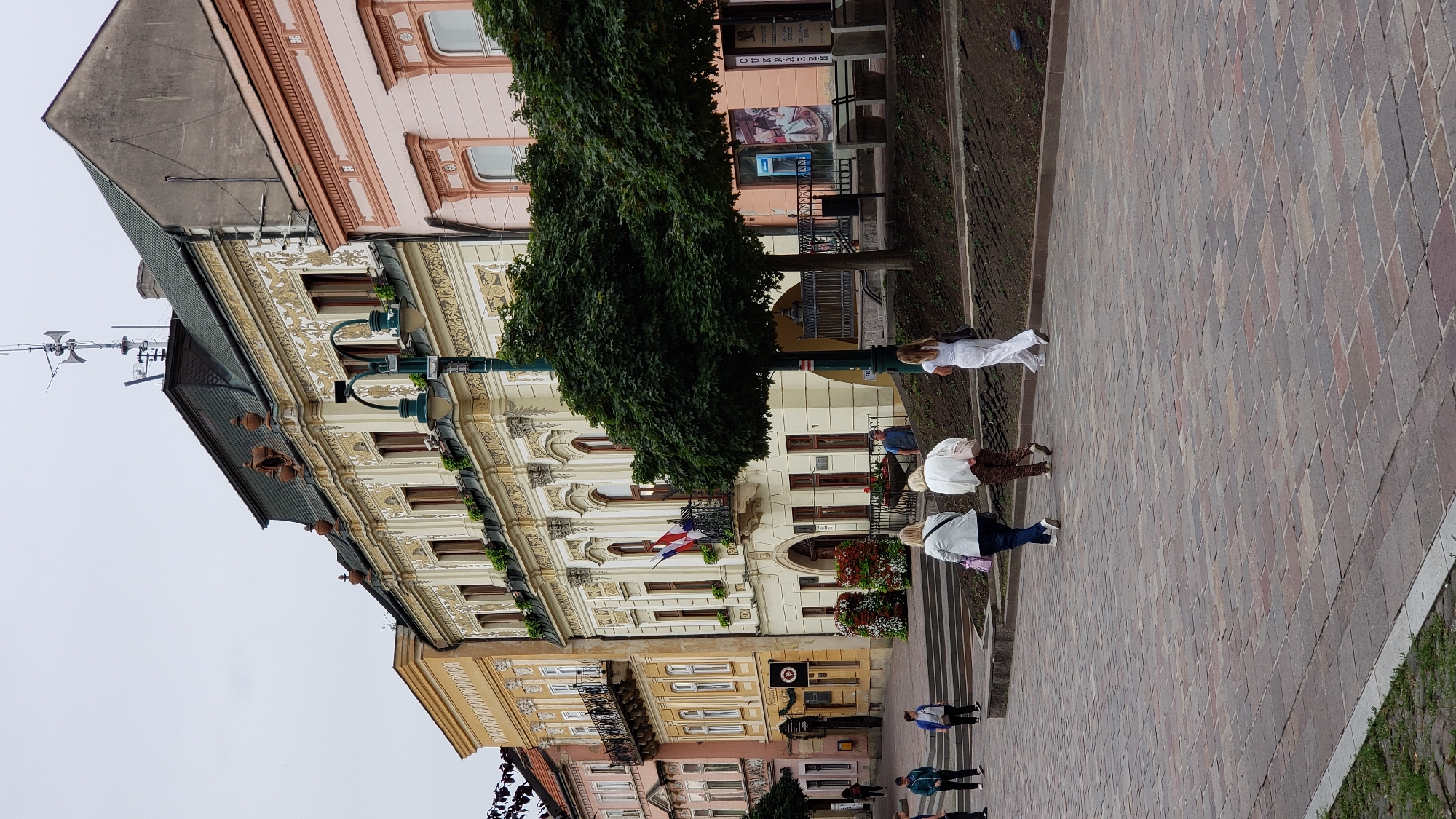 The image depicts a street scene in the city of Prešov, Slovakia. In the foreground, a paved walkway with a brick pattern stretches across the frame. Several individuals are walking on this walkway. In the middle ground, a tiered planter area with greenery and flower arrangements is visible, separating the walkway from a raised sidewalk. Beyond the sidewalk, a row of buildings with ornate facades line the street. One building, prominently featured, is pale yellow with intricate decorative moldings and a dark green mansard roof. A flag is displayed on a balcony of this building. Adjacent to it are other buildings, one pink and another yellow, with various architectural details including windows and balconies. Trees with full green canopies are planted along the sidewalk. In the background, the sky is overcast.