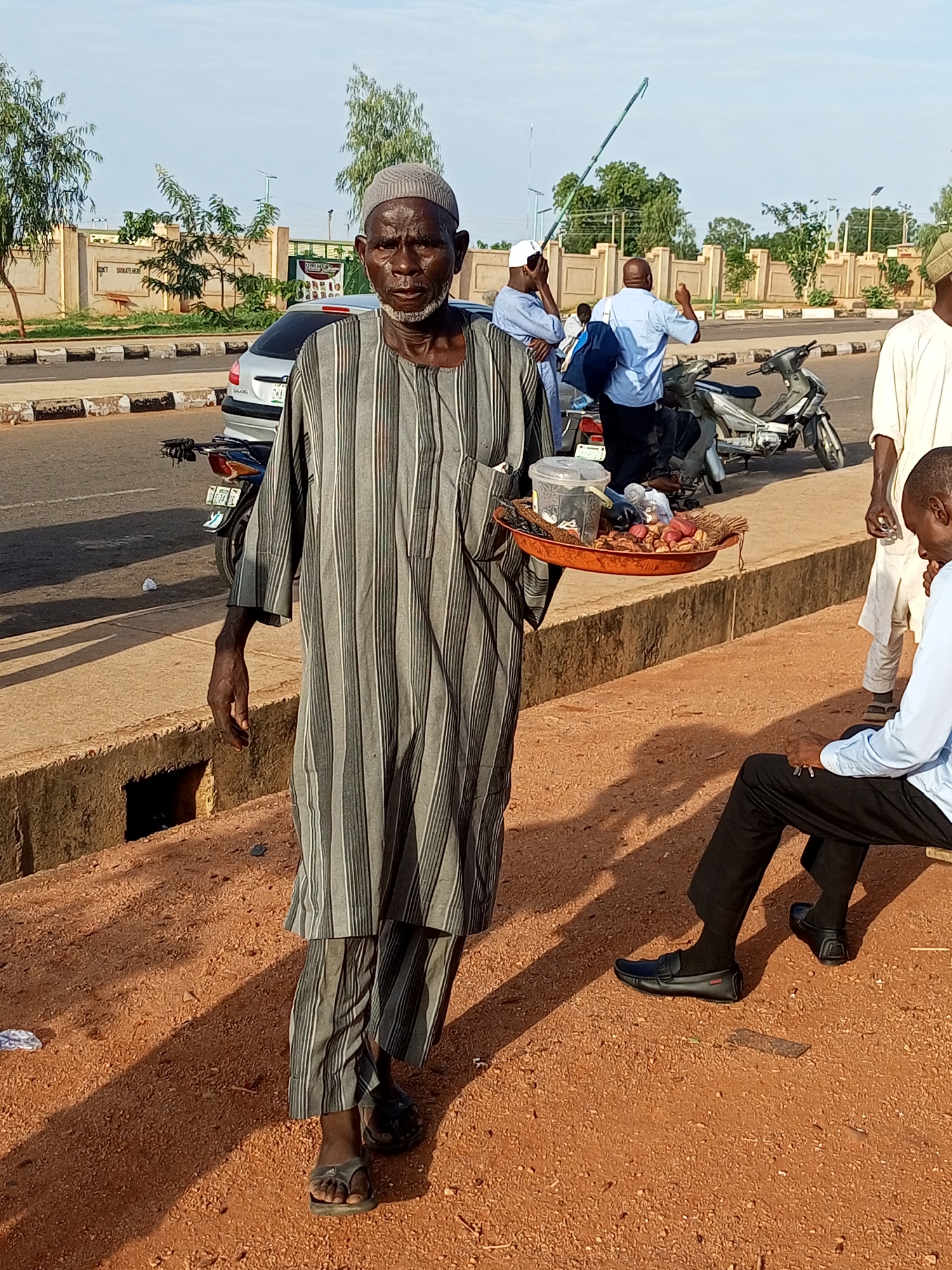In Gombe, Nigeria, an elderly man with a weathered face and a gray and white striped traditional outfit walks towards the camera, carrying a round tray filled with various food items, including what appear to be dates or dried fruit, and small containers of liquids or pastes. He wears a gray skullcap and dark sandals. The ground is covered in reddish-brown soil with sparse gravel. Behind him, a street is visible with a silver car and a motorcycle parked on the side. Other individuals, some in light blue and white attire, are present in the background, interacting or standing near the vehicles. A seated man in a light blue shirt and black pants is on the right side of the frame, his gaze directed downwards. The environment suggests an outdoor, potentially market or roadside setting, with a building facade and trees visible in the distance. The lighting indicates it is daytime, possibly late afternoon, given the long shadows cast on the ground.