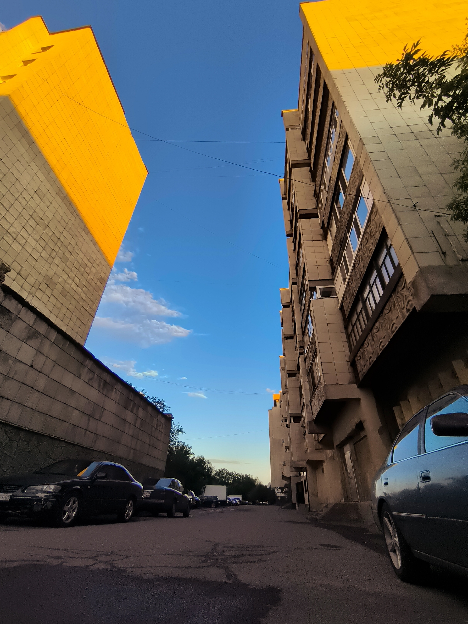 The image depicts a street scene in Qarağandı city administration, Kazakhstan, featuring an overcast sky and a mixture of residential and commercial buildings. The foreground is dominated by an asphalt road with visible cracks and puddles, indicating recent rain. Parked on the side of the road are several dark-colored cars, some with visible damage or wear.
To the left, a tall, beige building with a yellow upper section rises, its lower half covered by a textured concrete wall. The yellow section appears to be a contrasting architectural element, possibly a newer addition or a distinct design choice. Overhead, power lines crisscross the sky.
To the right, a multi-story building with a neutral facade, featuring multiple windows and decorative stone elements, stands prominently. The building's architecture suggests it is an older structure, possibly from the Soviet era, given the style of the windows and the building's overall form. A portion of a dark-colored car is visible in the lower right corner of the frame, its side reflecting the sky and surrounding buildings.
In the background, more buildings and trees are visible, contributing to the urban landscape. The overall impression is of a typical city street in Qarağandı, with elements of both older and newer construction. The lighting suggests it might be late afternoon or early evening, with the sky transitioning between day and night.