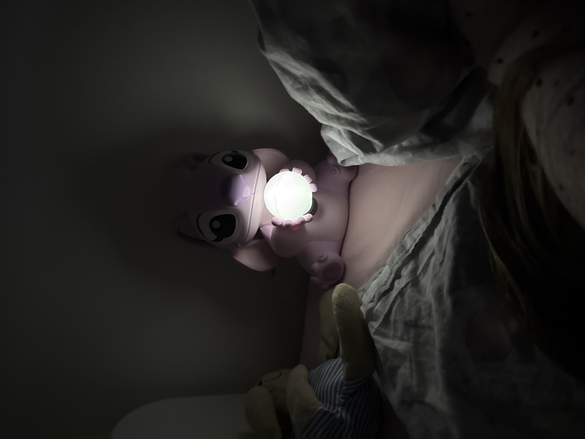In a dimly lit room, likely a bedroom, a pink "Angel" plush toy sits beside a person lying in bed. The plush toy holds a glowing white orb in its paws, illuminating its face. The person is partially covered by a grey and white striped blanket. A stuffed toy with light beige hands and a blue striped body sits below the "Angel" plush. The environment is dark, with the only light source being the orb held by the plush toy. The location cannot be determined to be Sofia, Bulgaria from the image alone.