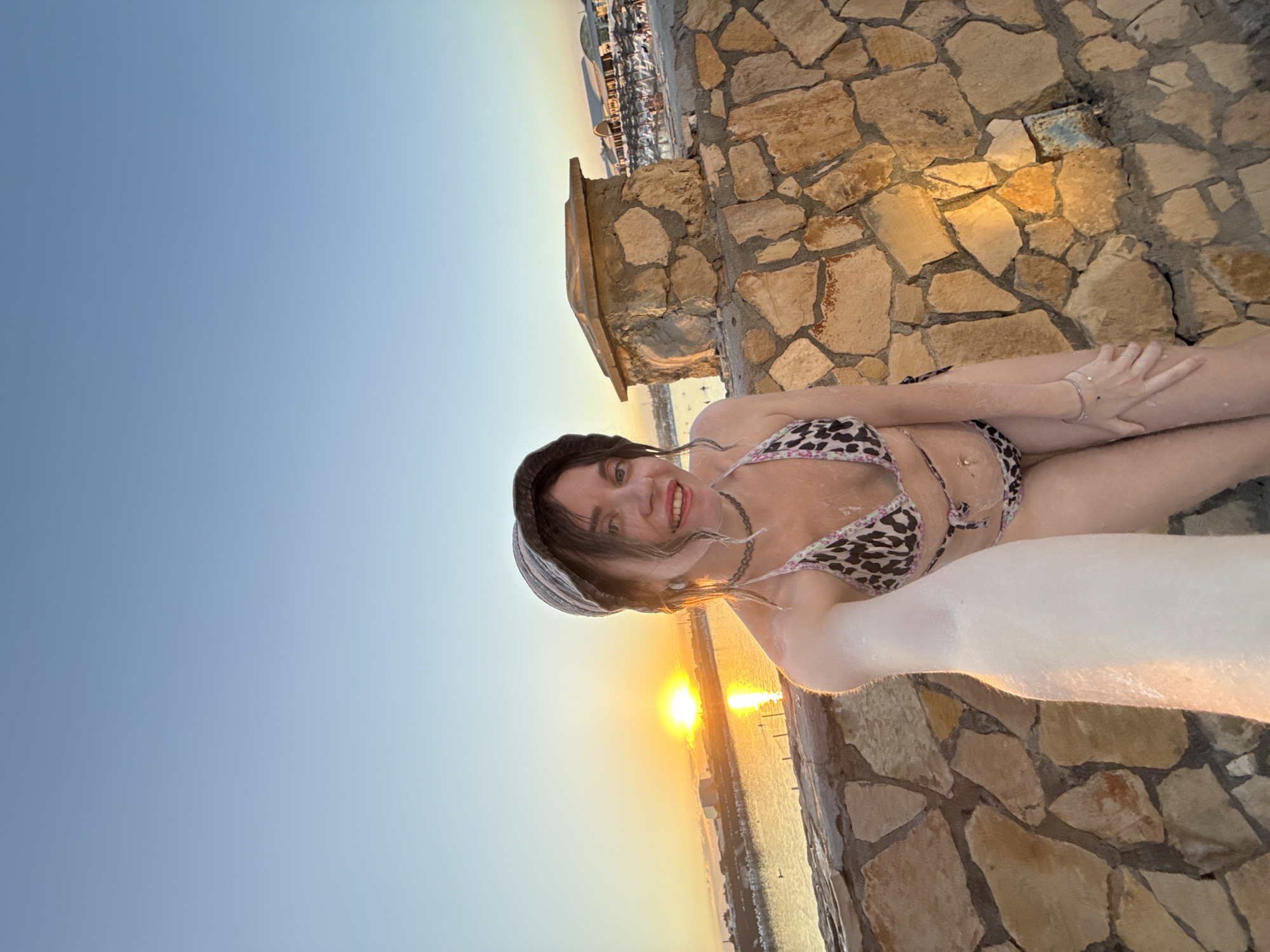 A woman in a leopard-print bikini and a bucket hat takes a selfie on a stone wall overlooking a body of water at sunset. The sun is low in the sky, casting a warm glow on the scene. In the background, there are buildings and boats, suggesting a harbor or a resort area. The woman has white powder on her skin and hair, possibly salt from a saline lake, which are common in Sol-Iletsk.