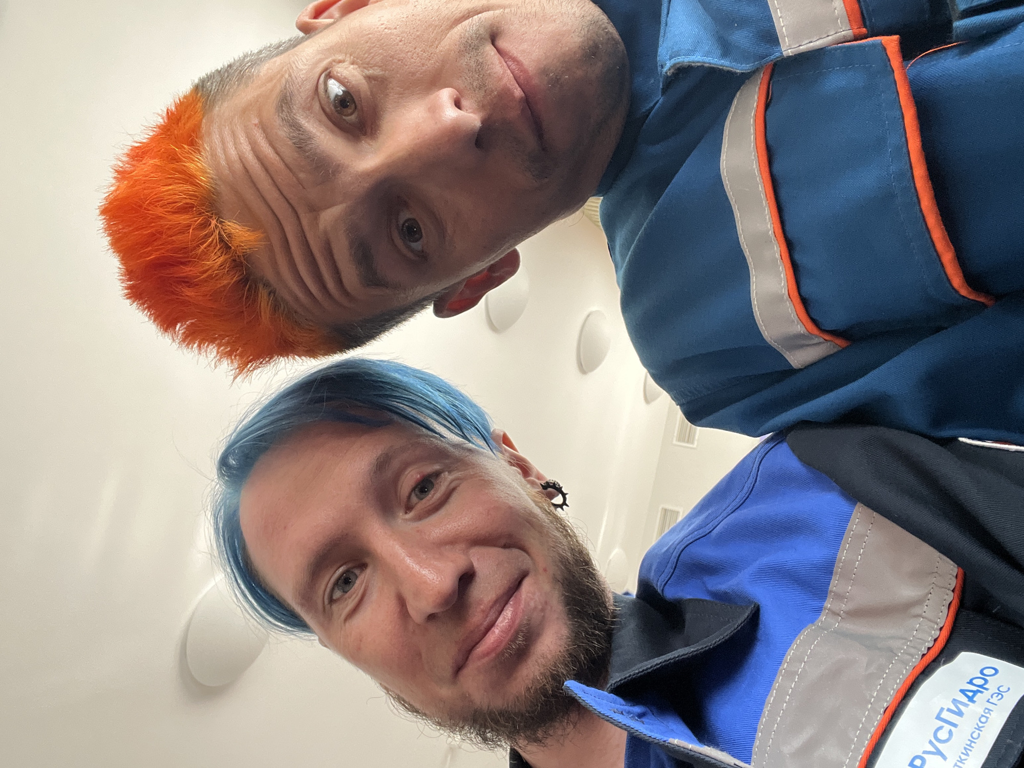 Two Caucasian males are captured in a close-up selfie, likely inside a building in the city of Chaikovsky, Russia. The male at the top of the frame has short orange hair and is wearing a blue uniform. The male at the bottom has blue hair, a beard, and an earring. He is also wearing a blue uniform, which features orange trim and reflective grey panels. A Russian logo is visible on the uniform. The background is a white ceiling with circular light fixtures.