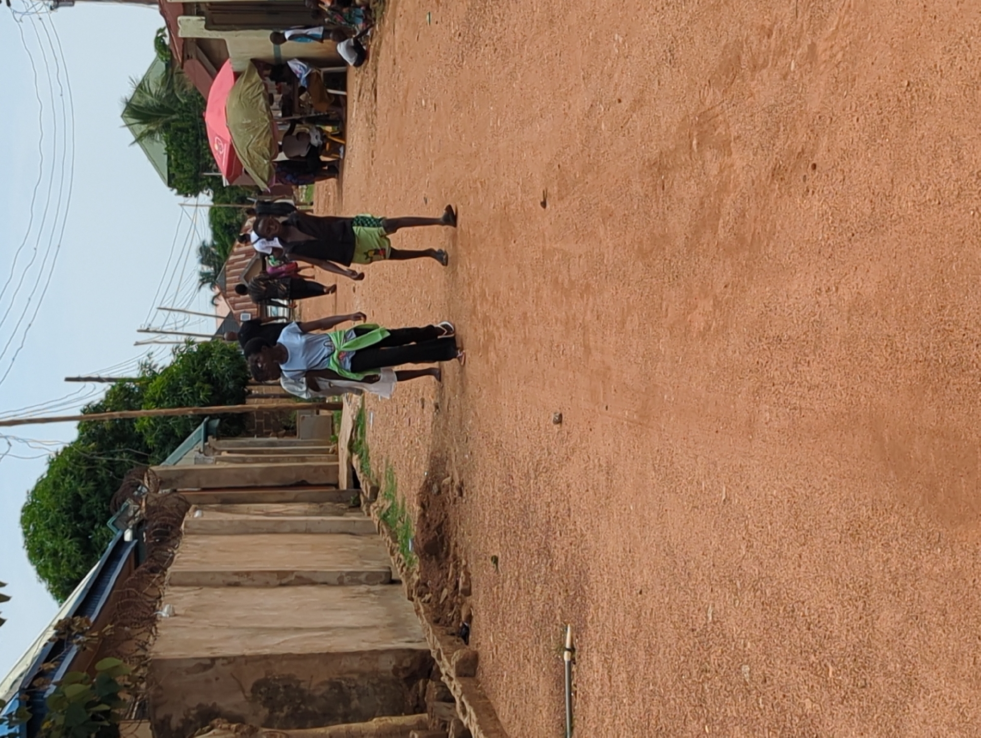 The image depicts a dusty, unpaved street in Sabon Tasha, Nigeria. Two people are walking on the road, one wearing a light-colored shirt with a green sweater tied around their shoulders and dark pants, and the other wearing a dark shirt and green patterned shorts. There are other people and parasols in the background. A set of concrete steps runs along the left side of the road. Barbed wire is installed on the house to the left. Powerlines can be seen overhead.