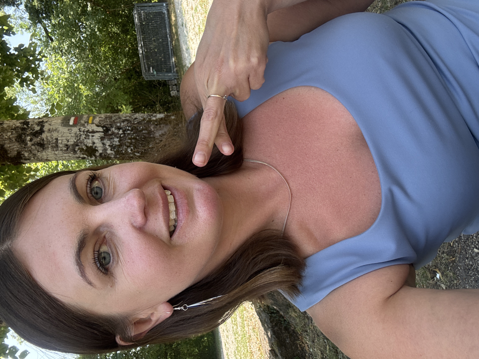 A woman with brown hair and blue eyes is taking a selfie outdoors. She is wearing a light blue dress and a silver necklace. Her right hand is raised, with two fingers extended, and she is wearing a ring on her right ring finger. She is smiling and looking at the camera. The background shows trees, foliage, and a tree with trail markers. In the distance, there is a metal fence. The scene appears to be a park or natural area.