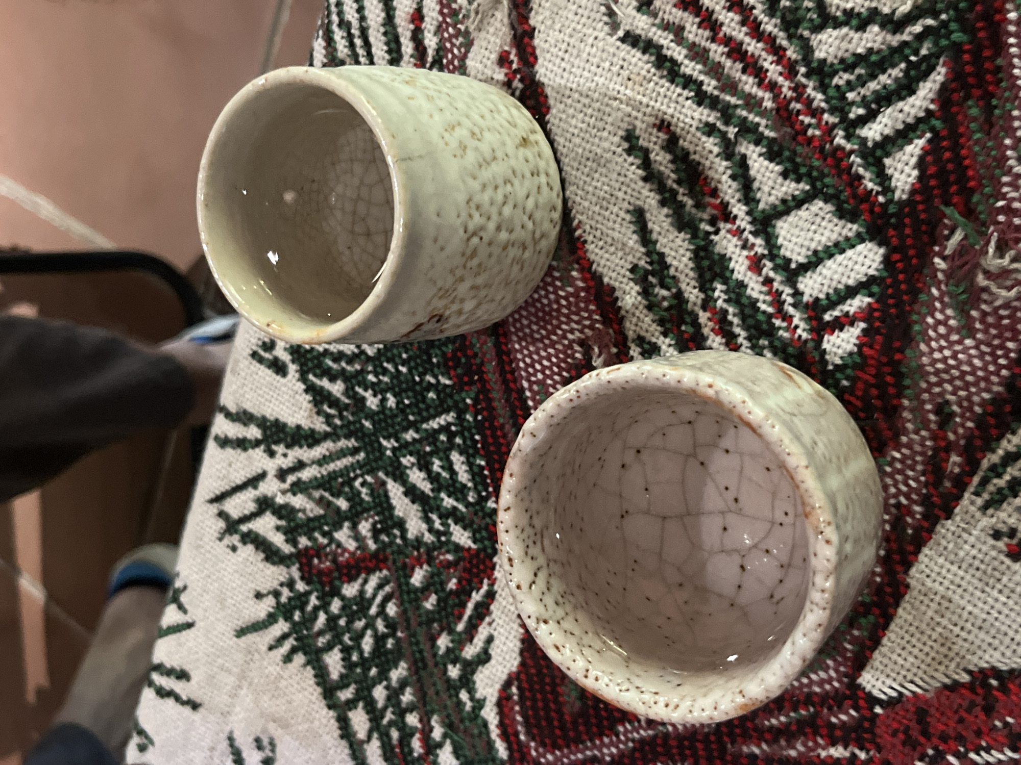 The image shows two ceramic cups filled with a light-colored liquid, likely sake, placed on a woven tablecloth with a festive design featuring green pine needles and red accents. The cups have a speckled, light beige exterior with a crackled glaze on the interior. One cup is positioned in the foreground, slightly to the left, while the other is to the right and further back, with its rim facing the camera. In the background, blurred elements suggest a person's legs and feet wearing dark pants and blue and white shoes, as well as a wooden floor or surface. The scene is lit with natural or overhead lighting. The location is not identifiable within the city of Đà Nẵng, Vietnam.