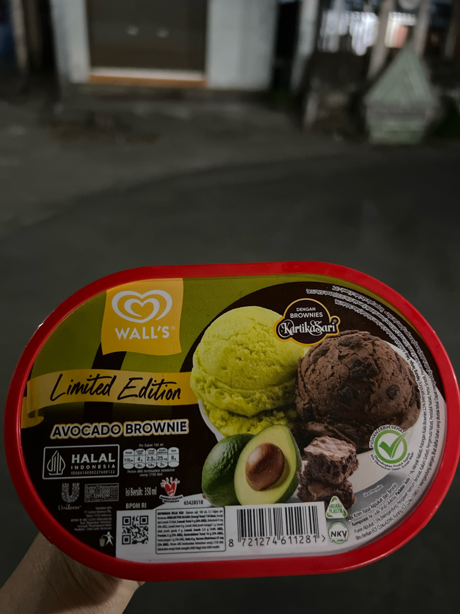 An outdoor, low-angle, medium shot captures a red-rimmed, oval tub of Wall's Avocado Brownie limited edition ice cream, held by a person's fingers. The tub is predominantly green, featuring images of avocado halves, chocolate brownies, and scoops of avocado and chocolate ice cream. Prominently displayed are the "Wall's" logo, "Limited Edition" text, and the product name "AVOCADO BROWNIE." Nutritional information and Indonesian Halal certification are visible. A barcode and QR code are printed on the tub. The background is blurred, showing a dimly lit, indistinct urban environment, possibly a street or pathway in Tlogoadi, Indonesia, with some structures and faint lighting, suggesting it's nighttime or dusk. The overall scene is dark and slightly grainy.