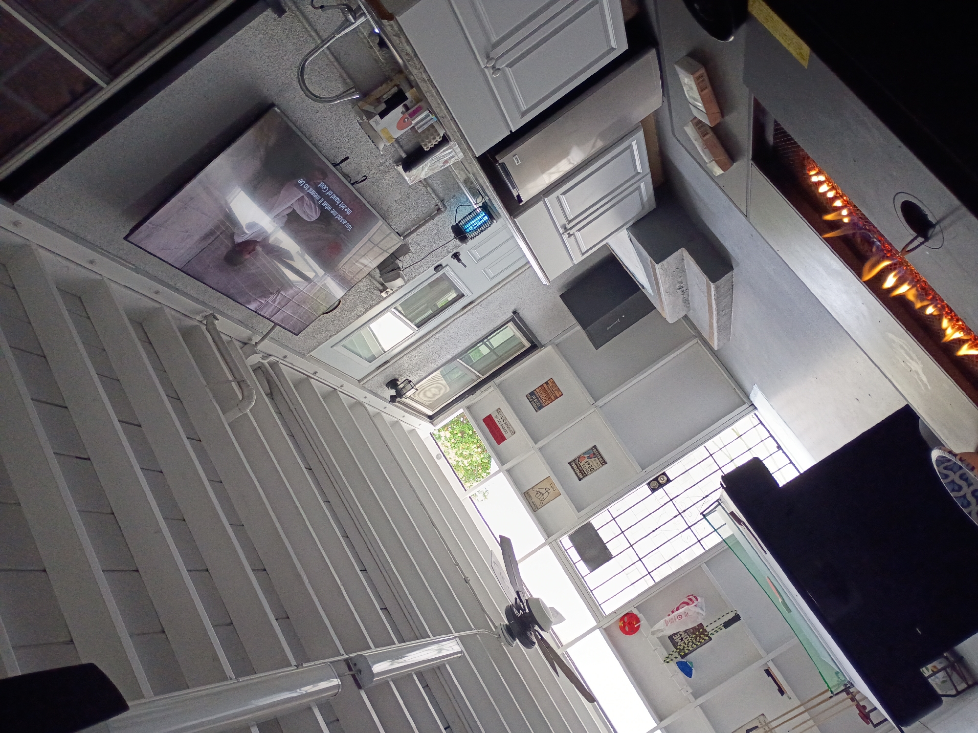 Here is a forensic description of the scene:
**General Description:**
The image shows an interior scene with an unusual perspective, tilted significantly. The space seems to be a combination of a kitchen/bar area and a more open area with what appears to be a partially covered patio or courtyard.
**Environment:**
* **Kitchen/Bar Area:** This area includes countertops, cabinets (white), a stainless-steel refrigerator, a sink, and a fireplace. A television is mounted above the countertop.
* **Open Area/Patio:** This area features a white slatted ceiling or roof structure, a ceiling fan, and what appears to be a large window or opening to the outside. There is an open area visible through the windows with greenery.
**Objects:**
* **Television:** Shows a scene with text; "You asked me what it meant to be the left hand of God" can be read.
* **Cabinets and Countertops:** Standard kitchen fixtures.
* **Ceiling Fan:** Located in the open area.
* **Fireplace:** Built into the countertop of the bar, with flames visible.
* **Assorted Items:** Including kitchen supplies, decorative items on shelves, and what might be advertising posters or signs.
* **Wall Decor:** Posters or signs with text and images, including the words "DEAF ENGLISH ONLY CUSTOMER BEHAVIOR DEPENDING ON"
**People:**
* The scene is devoid of people.
**Location:**
* Due to the architectural style and the signage, it is possible that this interior is located somewhere in the city of Los Angeles, United States of America.
**Interactions:**
* There are no interactions taking place in the scene.
**Overall Impression:**
The scene is somewhat cluttered with items and has an unusual perspective, which makes it more difficult to determine the layout and use of the space.
