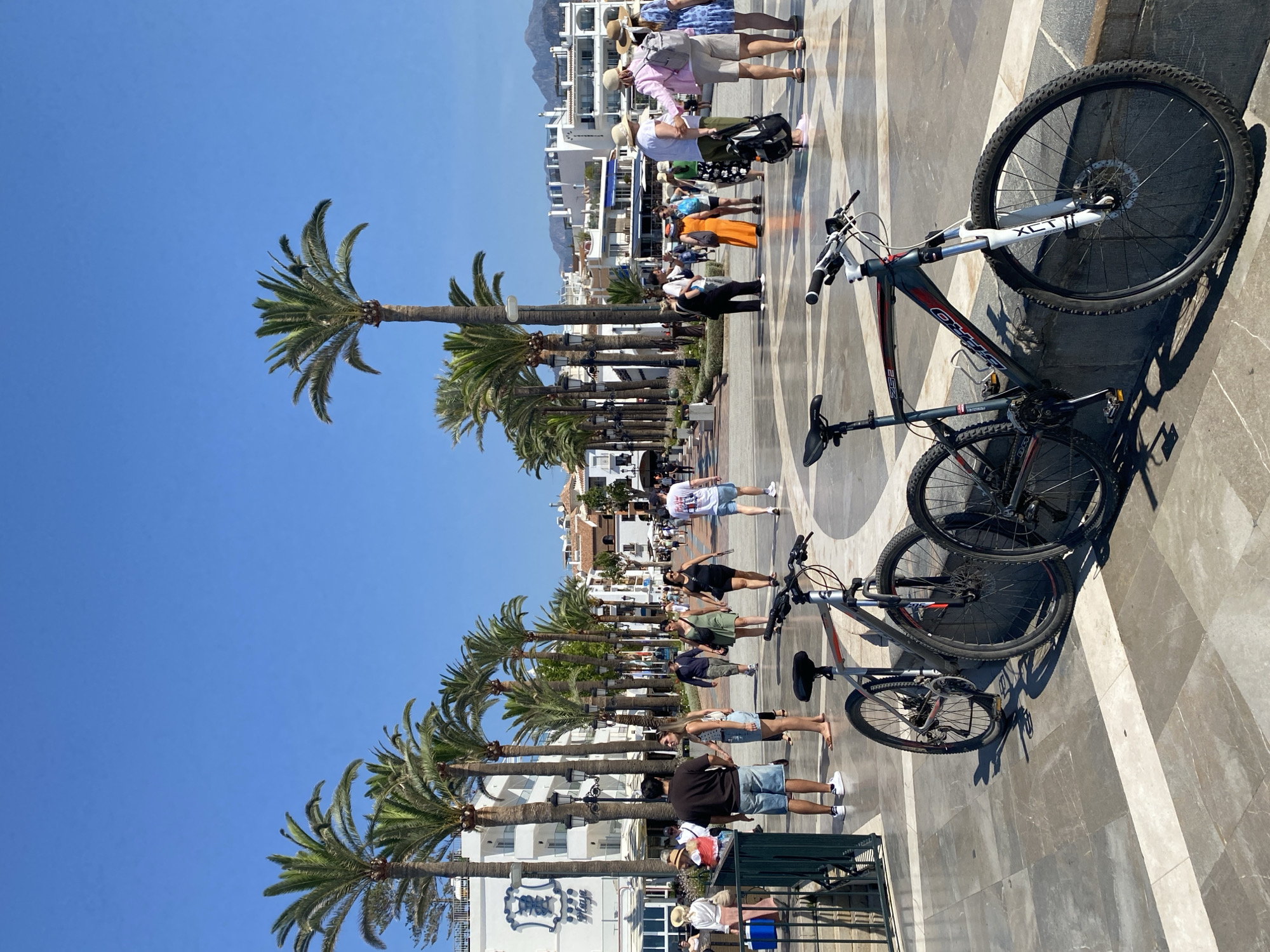 This is a ground-level, eye-level, medium shot photograph taken on a sunny day, likely in Nerja, Spain, given the context provided by the palm trees and the white Mediterranean-style buildings. The scene depicts a wide, paved promenade lined with numerous tall palm trees. In the foreground, two bicycles are parked on the promenade. The one on the left is a dark color with a red stripe on the frame and a silver front fork, while the one on the right is mostly silver and black. The promenade is paved with large, square, gray tiles interspersed with white borders.
In the mid-ground, a number of people are walking along the promenade. They are dressed in casual summer clothing, including shorts, t-shirts, dresses, and hats, suggesting a warm climate and a tourist destination. Some individuals are walking in pairs or small groups, while others are alone. The buildings on either side of the promenade appear to be hotels or shops, with white facades and balconies, some with blue awnings. The architecture is typical of coastal towns in southern Spain. In the background, a range of hazy mountains can be seen under a clear blue sky. The overall impression is one of a lively, popular public space on a bright, pleasant day.