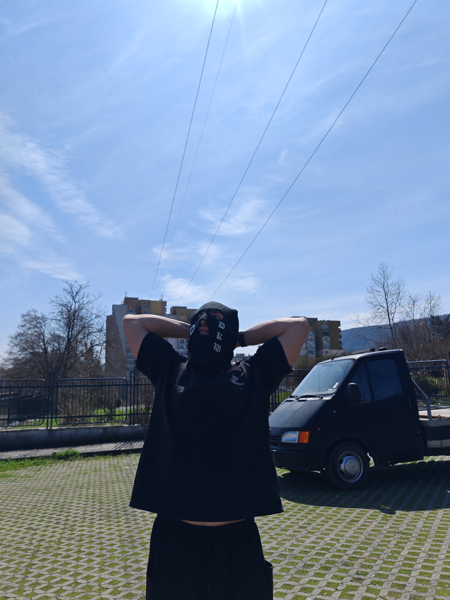 A daytime photograph depicts a single individual standing in an outdoor paved area within Shumen, Bulgaria. The individual, of unknown gender but appearing male, is positioned in the foreground, centrally, and faces the camera directly. They are wearing a black balaclava covering the entire face, with eyeholes visible and vertical white text, possibly "BLKW," present on the right cheek area. The individual is dressed in a black short-sleeved t-shirt and dark-colored trousers. Their hands are clasped behind the head, with elbows raised outwards.
To the immediate right of the individual, a dark-colored (likely black) flatbed tow truck is parked. The visible portion includes the front and side of the cab and a section of the flatbed, consistent with a Ford Transit model. The ground beneath the individual and the truck is a permeable pavement system composed of concrete grid pavers with green grass growing through the interstices.
In the mid-ground, a dark metal fence extends horizontally. Beyond this fence, to the left, a bare deciduous tree stands. Multiple multi-story residential buildings, identified as apartment blocks with varying facades (light-colored, yellow/orange, and grey), are visible in the background, characteristic of urban construction. A distant, tree-covered hill or low mountain range is present on the right horizon, typical of Shumen's landscape. The sky is light blue with sparse, wispy white clouds, and the sun is visible in the upper left quadrant, causing lens flare. Several parallel utility or power lines traverse the upper portion of the frame. The scene is consistent with an urban residential area near green spaces, situated at the base of the Shumen Plateau.