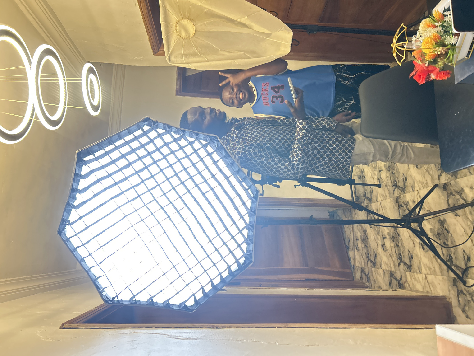 The image shows two individuals indoors, likely in a residential setting, as evidenced by the marble tiled floor. One individual is wearing a patterned shirt and light-colored pants, posing near a large octagonal softbox on a stand. The other is wearing a blue basketball jersey with the number 34 and the word "Bucks" visible, holding a phone and making a peace sign. In the background are a soft light umbrella and a table adorned with artificial flowers. Above, modern ring-shaped lighting fixtures are visible. The location is Unknown, Unknown.