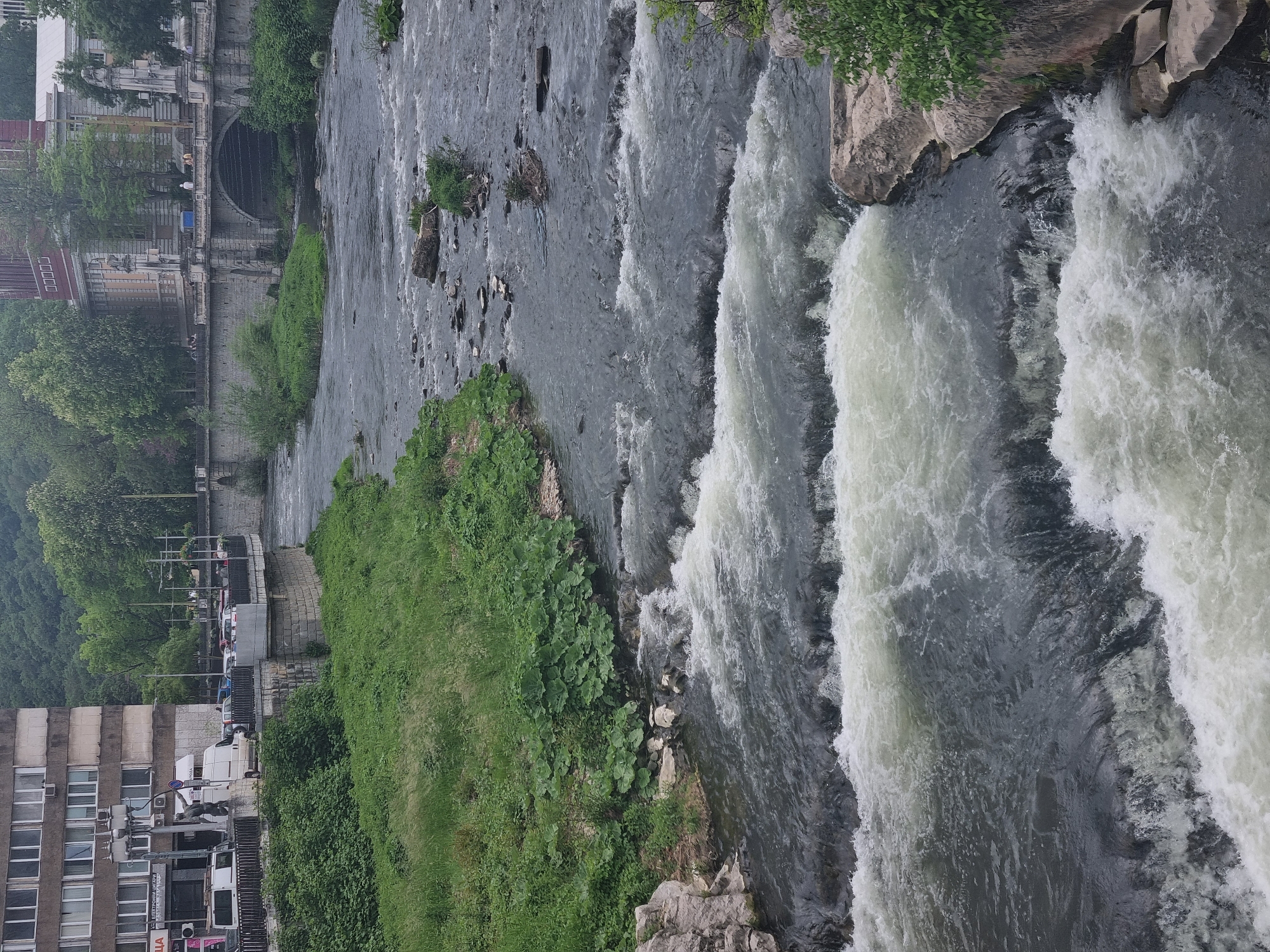 The image shows a view of the Yantra River in Gabrovo, Bulgaria. Fast-flowing water cascades over small waterfalls. The river is flanked by urban infrastructure, including buildings and a road with parked vehicles on one side, and vegetation on both sides. A bridge with a tunnel can be seen further upstream.