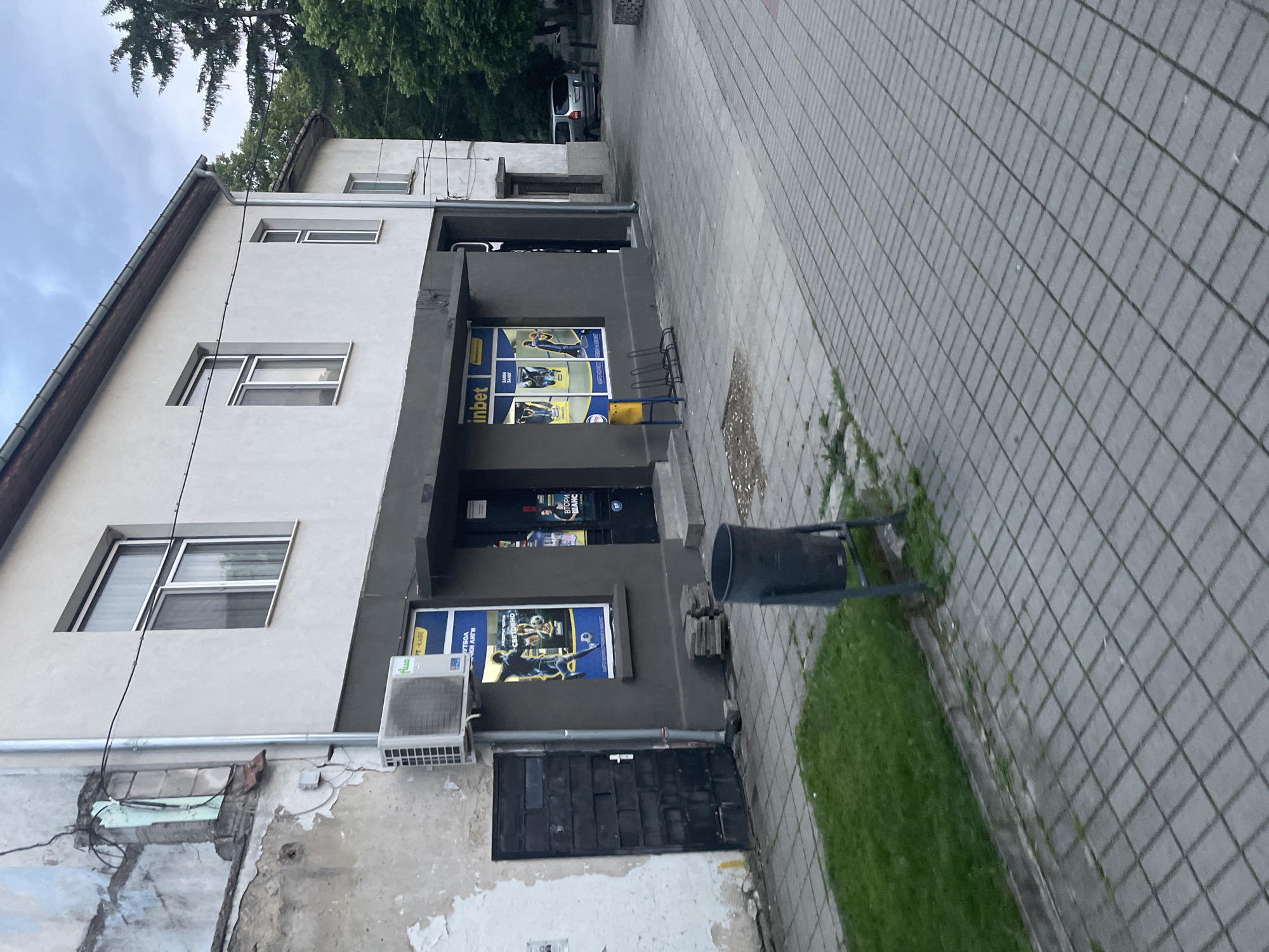 The image displays a quiet street scene in Ihtiman, Bulgaria, during what appears to be an overcast daytime. The primary subject is a two-story building with a multi-toned facade. The ground floor is painted a dark gray, housing what appears to be a commercial establishment, possibly a betting shop. Prominent blue and yellow advertising posters are displayed in the windows and on the wall, featuring the brand "inbet" and various sports-related imagery, including football players. Visible Bulgarian text on the posters includes "ФУТБОЛ" (football), "СВЕЩЕННО" (sacred/holy), and "ВТОРИ ШАНС" (second chance). An air conditioning unit with the brand "FLUO" is mounted on the wall below one of the posters. A black, metal door is situated at the bottom left of the ground floor. An empty black bike rack is positioned near the entrance.
The upper story of the building has a lighter, off-white or light beige facade, featuring multiple rectangular windows with white frames. A pitched roof with a dark gutter system is visible against an overcast sky and green trees in the background.
To the right of the building, a wide paved walkway made of rectangular gray tiles extends into the distance. A narrow strip of green grass separates the walkway from the building in some sections, alongside which stands a black cylindrical trash can. In the deeper background, lush green trees and a light-colored car (possibly white or silver) are visible. No people are present, and no particular activity is taking place, creating a static urban snapshot.