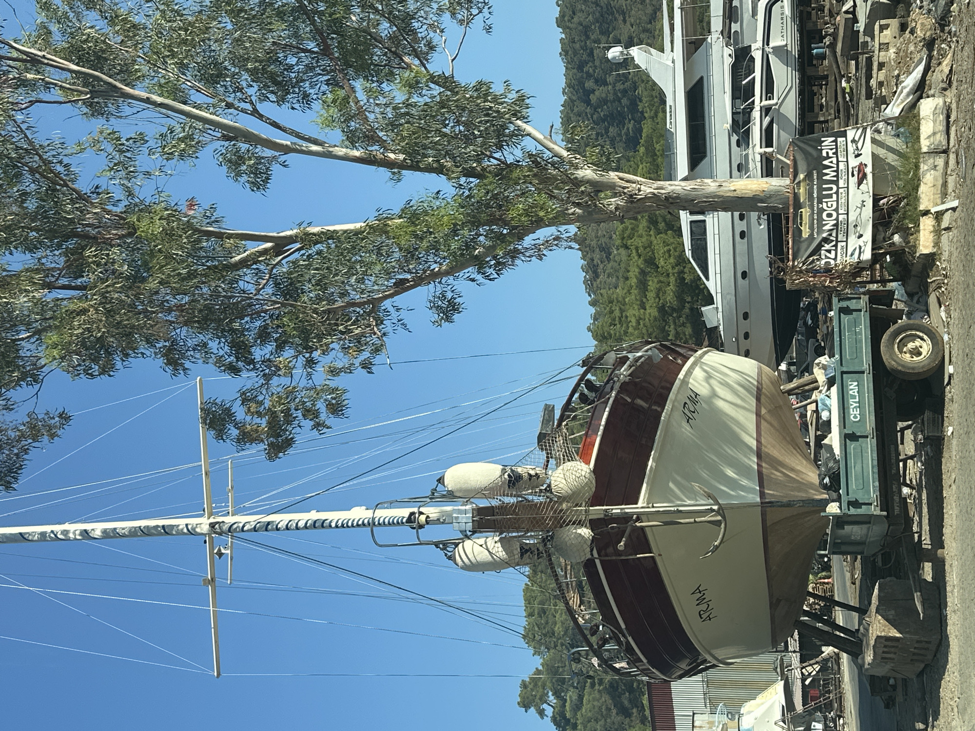 A sailboat named "ARMA" is dry-docked, supported by wooden stands, in an outdoor boatyard. The boat is painted with a dark red upper hull and a cream-colored lower hull, with the name "ARMA" clearly visible on both sides. It has a tall mast with numerous rigging lines. In the background, several other boats, including a large white yacht and smaller speedboats, are also visible. A tall eucalyptus tree with sparse green leaves stands to the right of the sailboat. The background shows a green, tree-covered hillside under a clear blue sky. A sign on the ground in front of the large yacht reads "OZKANOĞLU MARIN" and lists various services in Turkish. A green trailer with the word "CEYLAN" painted on it is positioned in front of the "ARMA" sailboat. The boatyard appears to be located in a coastal area, likely Fethiye, Turkey, given the presence of a marina and the style of the boats. There are no people visible in the scene.