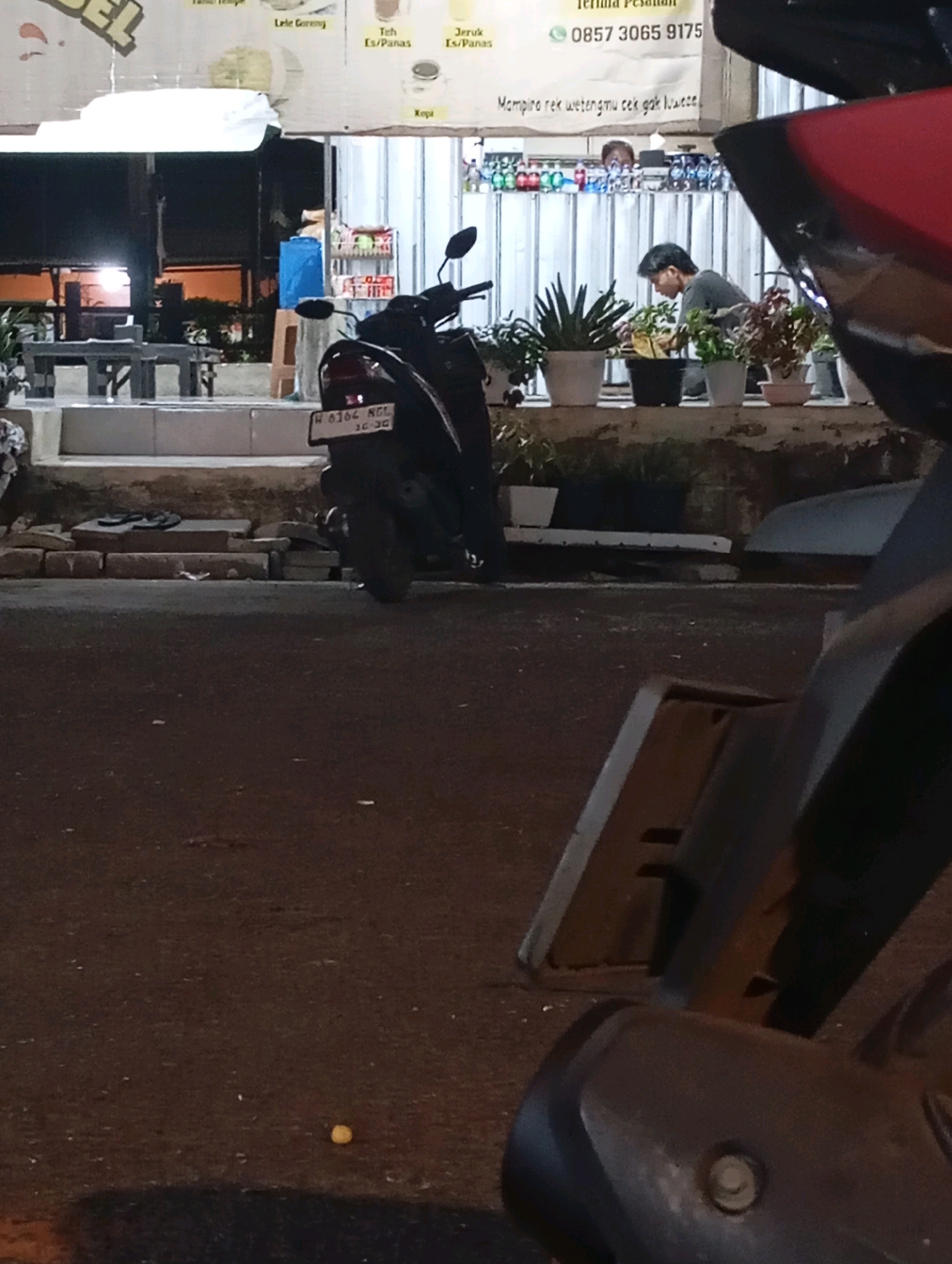 The image depicts a street scene at night in Randegan, Indonesia. In the foreground, a black motorcycle is parked, with its rear visible. The motorcycle has a license plate that reads "H 6164 NGL 10-20". Further back, a person, possibly a man with short dark hair, is tending to plants in pots arranged on a ledge. Behind this person, a row of bottled drinks is displayed on shelves. To the left of the plants and drinks, a blue container is visible on a stool. The background features a dimly lit establishment with what appears to be tables and chairs. The ground in front of the establishment is paved with bricks.