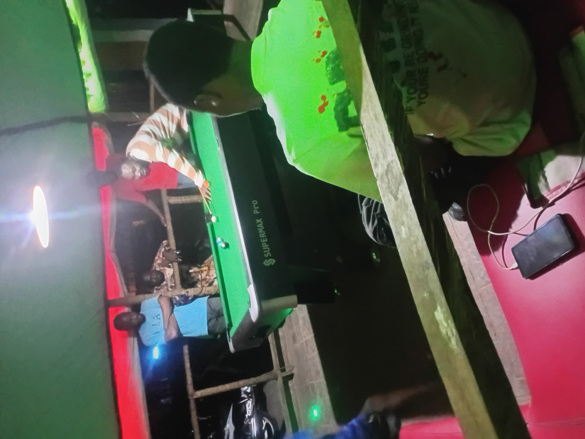 The image captures a lively evening scene in what appears to be a covered outdoor or semi-enclosed recreational area in Lokoja, Nigeria, possibly a local pool hall or a casual social venue. The primary activity is a game of pool, with several people gathered around the table.
In the foreground, the back of a person wearing a bright green t-shirt is prominent. Partially visible text on the shirt reads: "IF YOUR RE CRINDING YOU'RE DOING IT". This individual observes the game, positioned near a pool table. Another person, dressed in a light-colored striped shirt, is actively playing pool, leaning over the green-felt table with a cue stick. A few pool balls are visible on the table surface. The pool table itself is branded "SUPERMAX Pro".
Other individuals are present in the background: one person in a blue t-shirt stands by wooden support beams, and another can be partially seen further back. The setting features rustic wooden posts and beams, contributing to a makeshift or informal structure. A bright, circular light source illuminates the left side of the scene. On the right, a vibrant red surface, possibly a wall or a bench, holds a black smartphone that appears to be charging. Green fabric, resembling a tent or canopy material, is visible along the edges of the frame, providing cover. The dim ambient light and reliance on artificial illumination indicate that the scene takes place at night or in the evening, suggesting a relaxed social gathering.