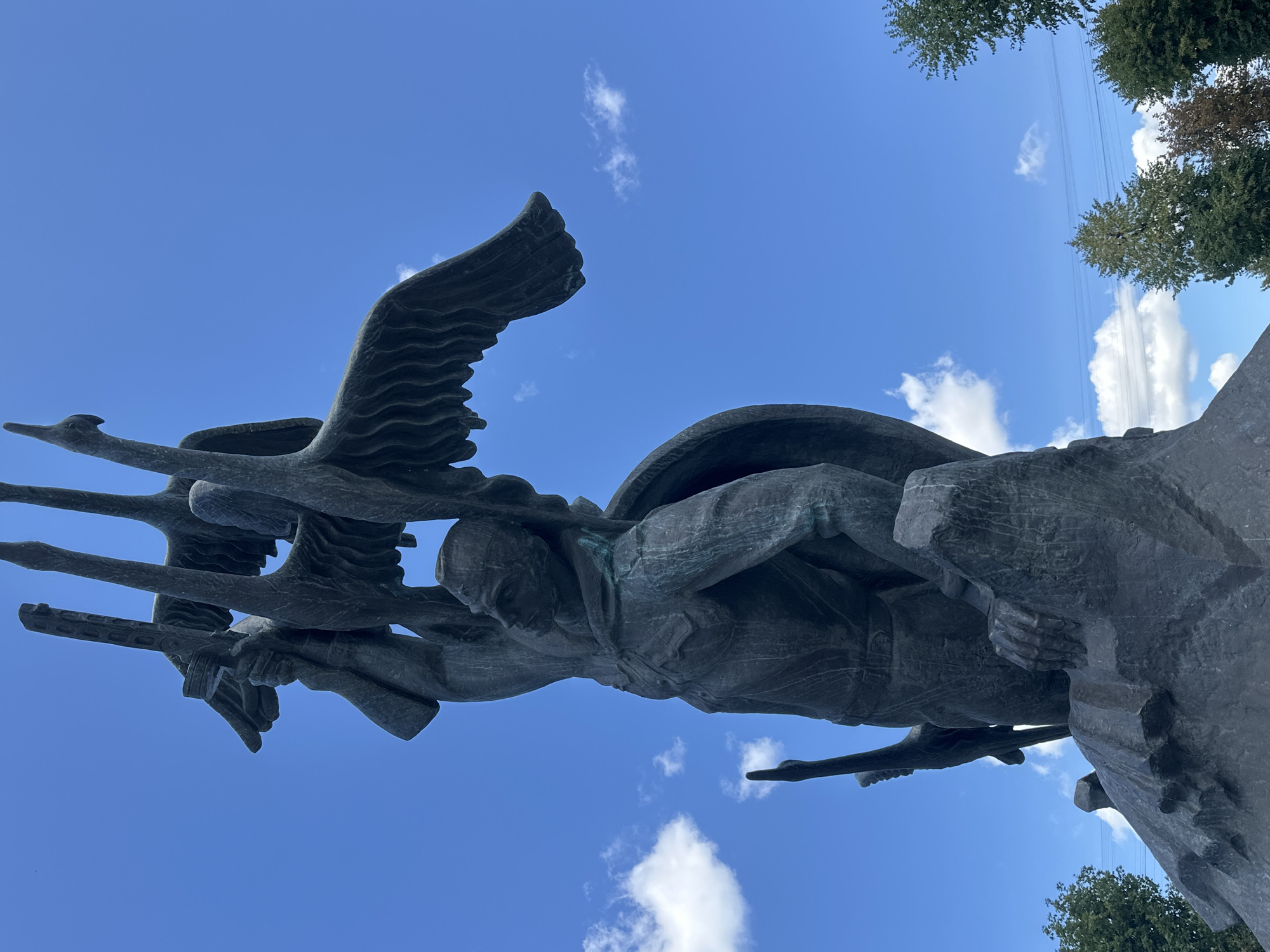 The image shows a bronze statue depicting a soldier holding up three cranes, symbolizing the fallen soldiers in the village of Zhovte, Ukraine. The statue stands on a rocky base against a clear blue sky with scattered white clouds. The soldier is dressed in military attire and appears to be in a determined pose, with one arm raised high and grasping the cranes. The cranes are depicted with their wings spread, as if in flight, and their long necks are held high. The statue is weathered, with a patina of green and brown, indicating its age. The background reveals a few trees on the right side of the frame. The overall impression is one of remembrance and solemnity.