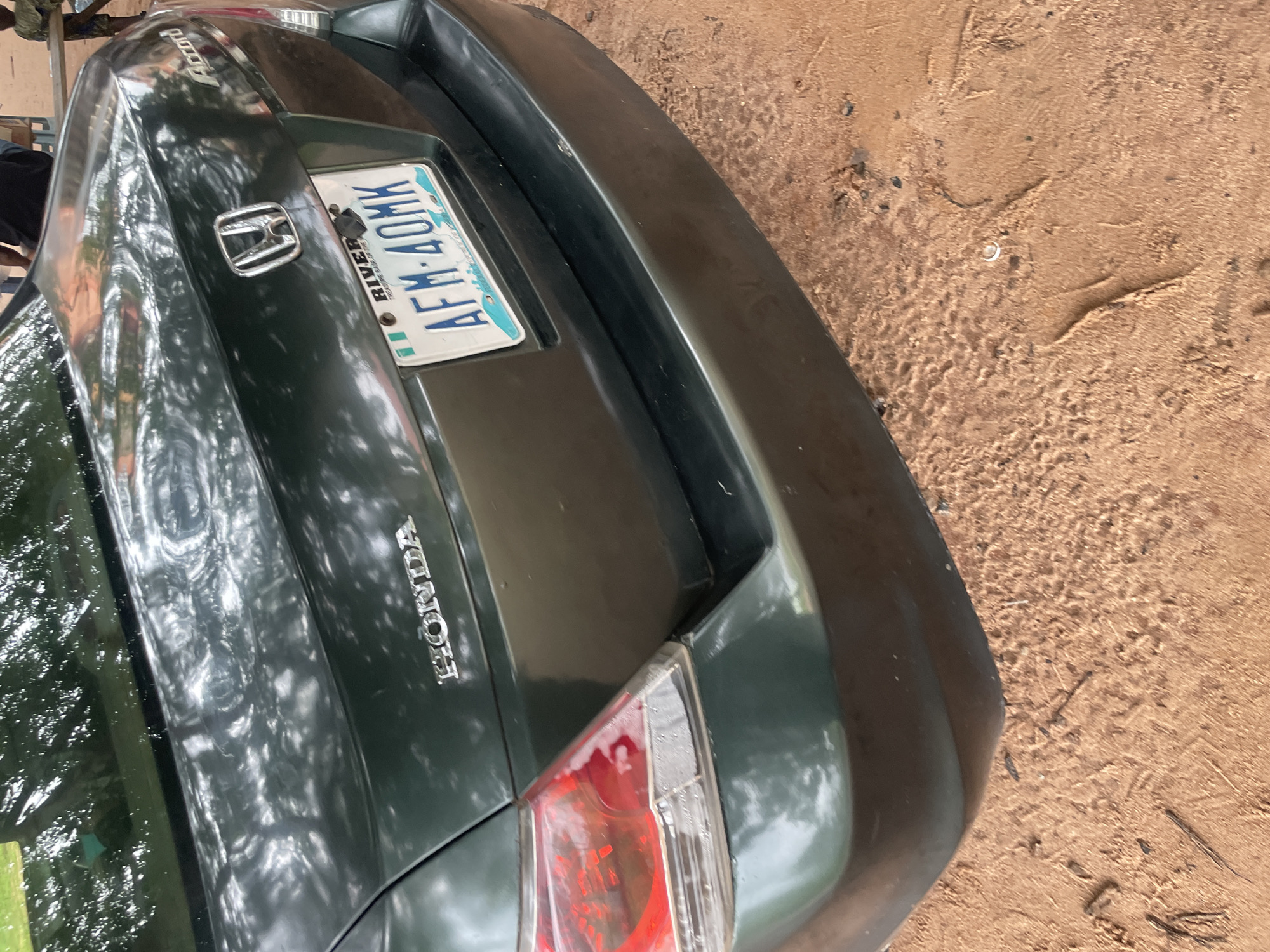 The image shows the rear passenger side of a dark green Honda Accord parked on a dirt road. The car's taillight and bumper are visible. The license plate reads "AFM-40MK" and has "RIVER STATE" printed on it. The car is reflecting dappled sunlight from trees overhead. The ground appears to be sandy or loamy soil, consistent with an outdoor environment. In the background, partially visible, are two individuals standing, one wearing a dark shirt and the other in a patterned shirt. A wooden structure is also discernible in the far background. The scene appears to be in Ihiagwa, Nigeria, as suggested by the location information.