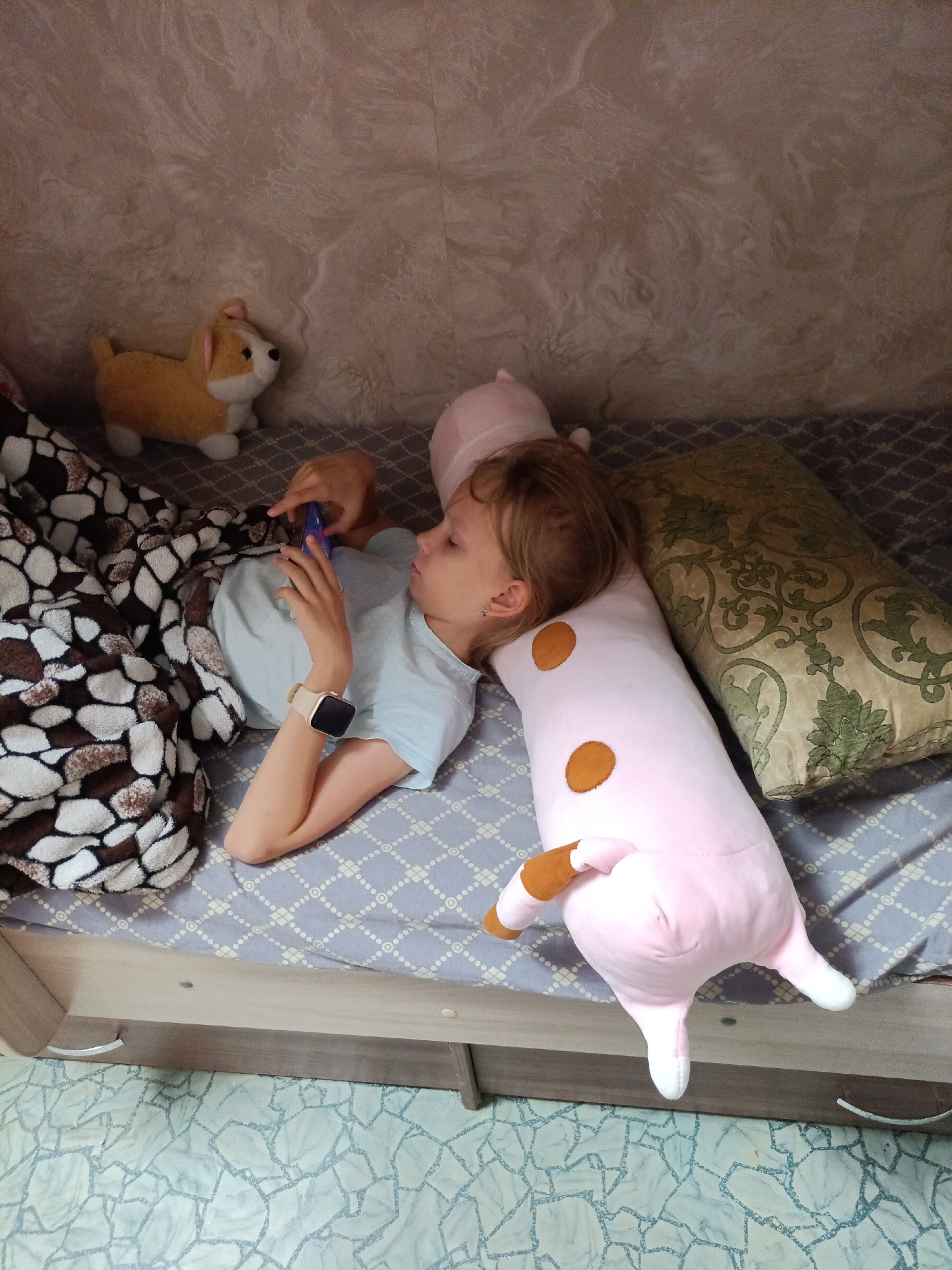 The image shows a young girl lying on a bed, looking at her phone. She is covered by a blanket with a brown and white patterned design. Beside her is a long, pink stuffed animal with brown spots and white paws. A decorative pillow with a green floral pattern is also on the bed. In the background, a small stuffed dog toy is visible on a textured wall. The bed appears to be in a bedroom. There is no specific indication of the city of Nyagan, Russia, in the image.