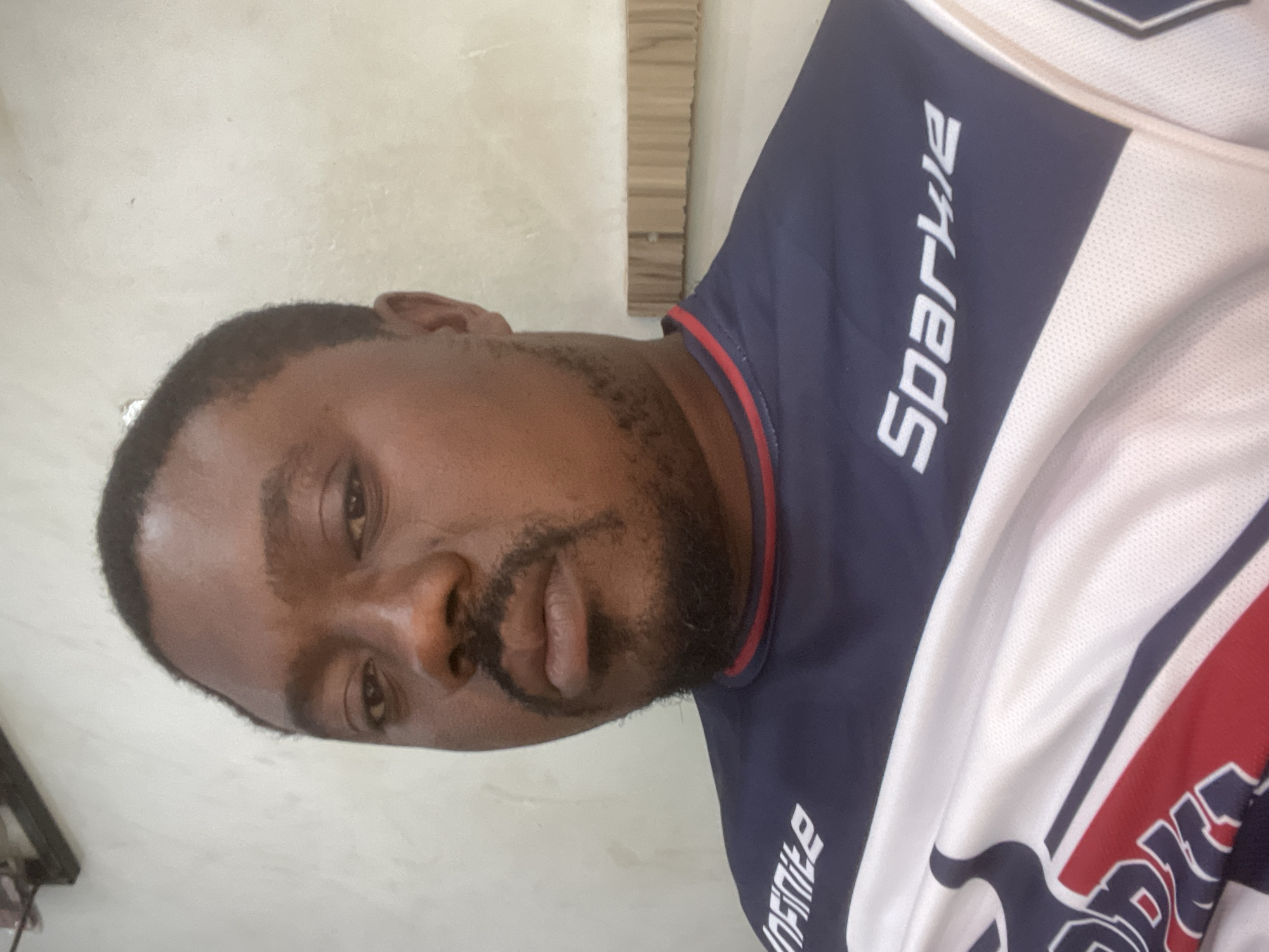 A man with dark skin, short black hair, and a beard is wearing a jersey with "Sparkle" written on the chest. The jersey is navy blue and white with red accents. The background appears to be an indoor space with light-colored walls and what looks like part of a wooden structure. There is no visible information to confirm this is in Jos, Nigeria.