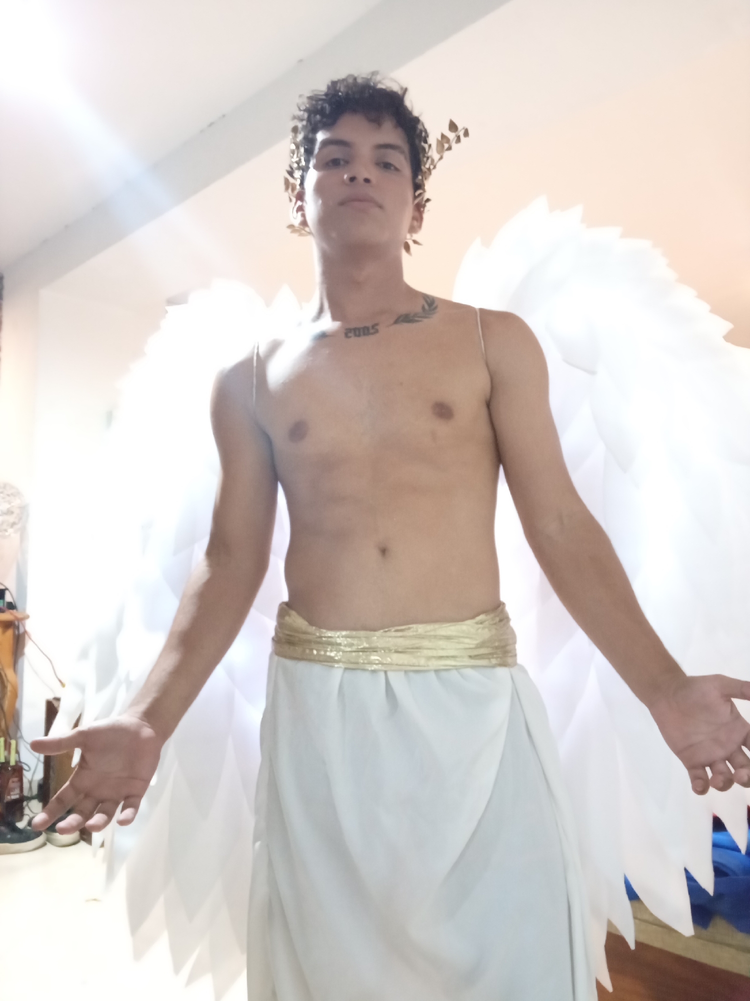 A young man stands indoors, dressed as an angel with large white wings and a gold laurel wreath on his head. He is shirtless, wearing a white garment cinched with a gold belt. The setting is not discernible as Playa El Agua, Venezuela based on the background details, which show a simple interior setting. The person's arms are extended slightly outwards. The lighting is somewhat bright, possibly from indoor lights or a nearby window. A tattoo is visible on his upper chest.