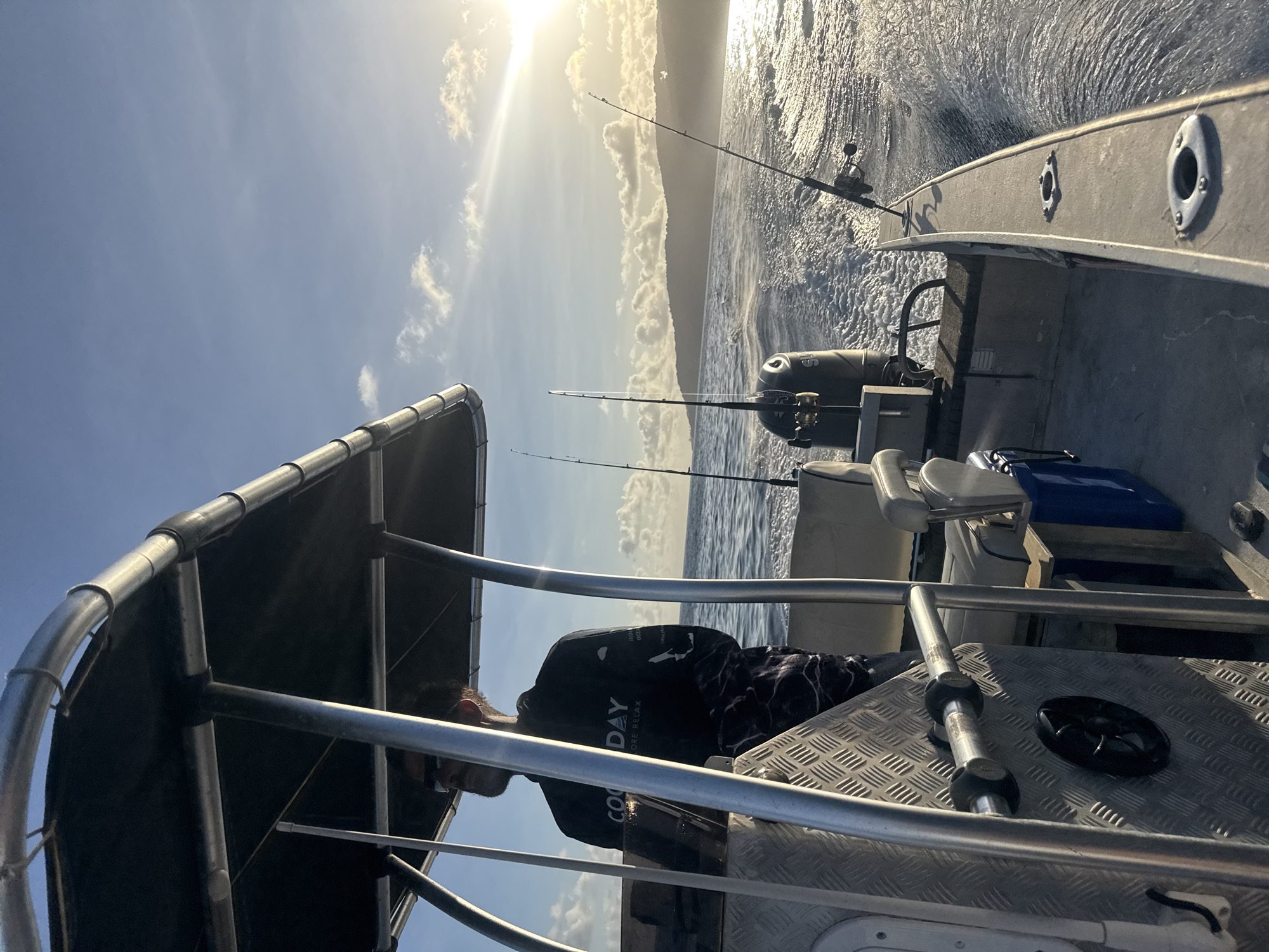 A person is driving a boat with a canopy, wearing sunglasses and a black long-sleeved shirt with camouflage pants. The boat is moving through the ocean, creating a wake. Several fishing rods are visible on the boat. The sun is setting in the background, casting a warm glow on the water and clouds. The location is Christmas Island, Christmas Island.