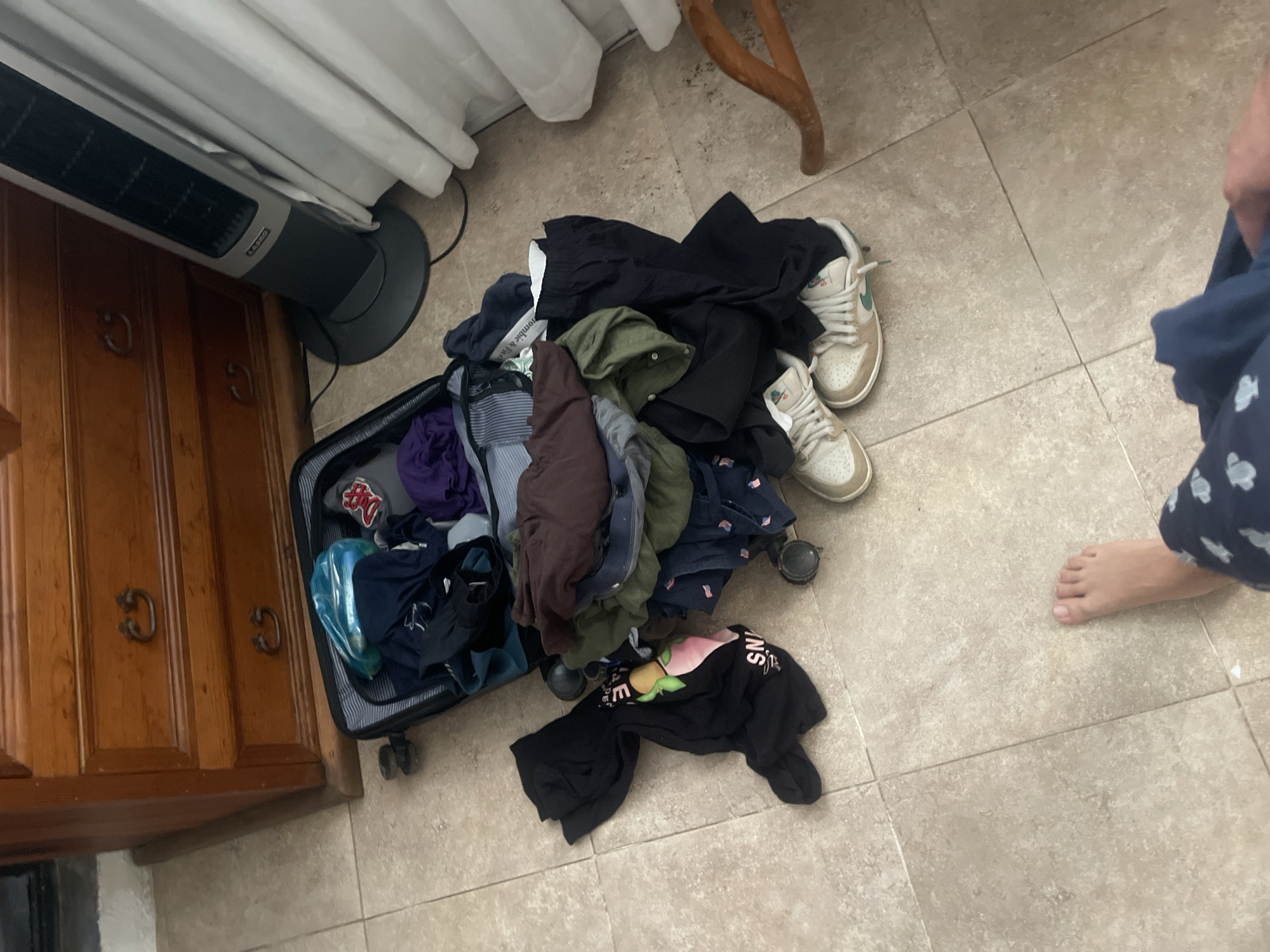 The image shows an open black suitcase on a tiled floor near a wooden cabinet in Oaxaca City, Mexico. The suitcase is overflowing with clothes of various colors and textures. Next to the suitcase is a pair of off-white sneakers. A partially visible person wearing blue patterned pajamas stands barefoot beside the clothes. A white curtain hangs in the background, next to a brown wooden chair leg. An upright Lasko fan is near the cabinet.