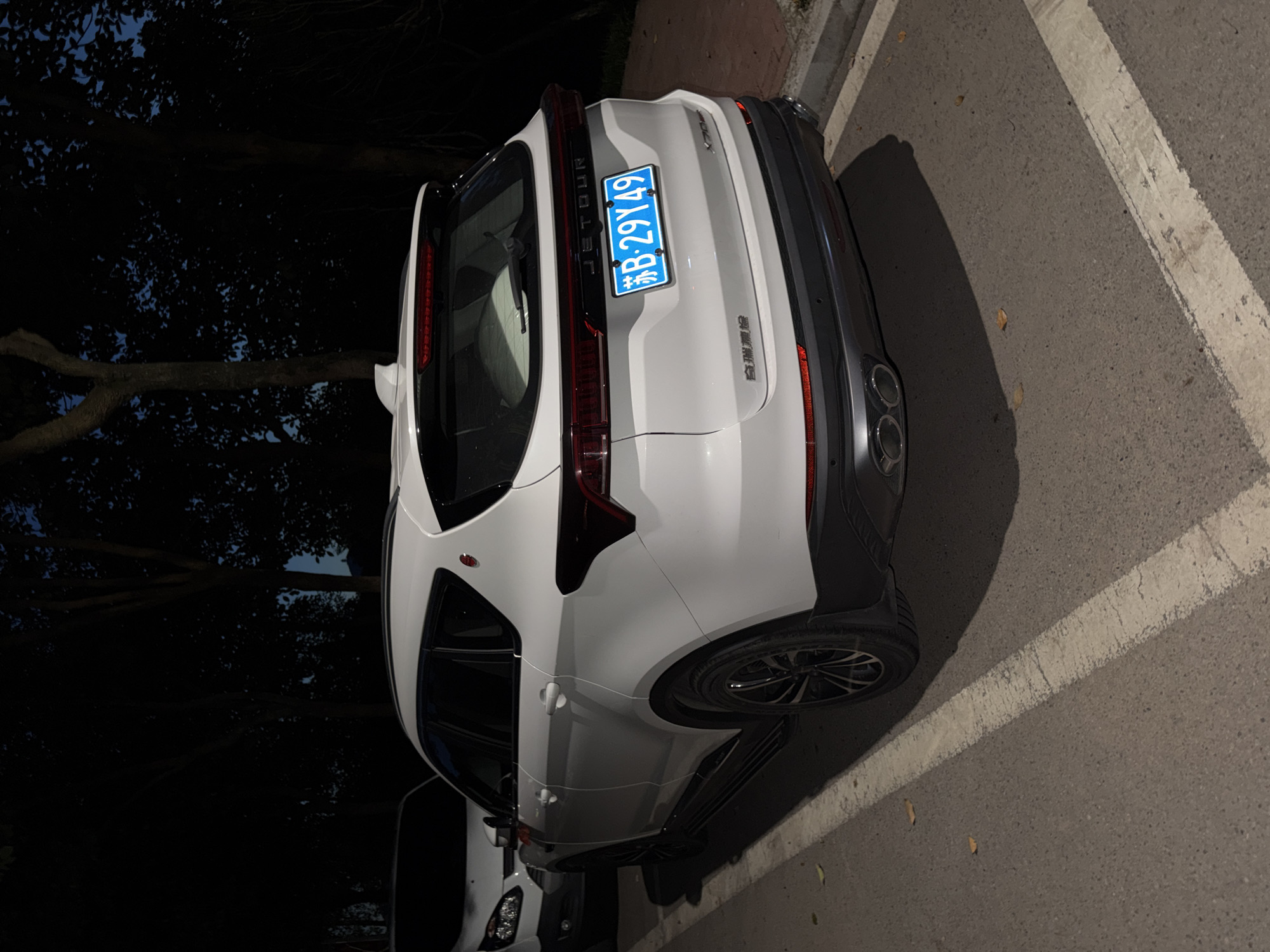 A white Jetour X70 Plus is parked on a paved road next to a curb. The car's rear is facing the camera, and the license plate is visible, reading "苏B 29Y49". The car is parked in a lot with a white line marking the parking space. In the background, trees with dark foliage are silhouetted against a dark sky. Another white vehicle is partially visible to the left of the Jetour. The scene appears to be in the city of Jiangjin, China, as indicated by the license plate.