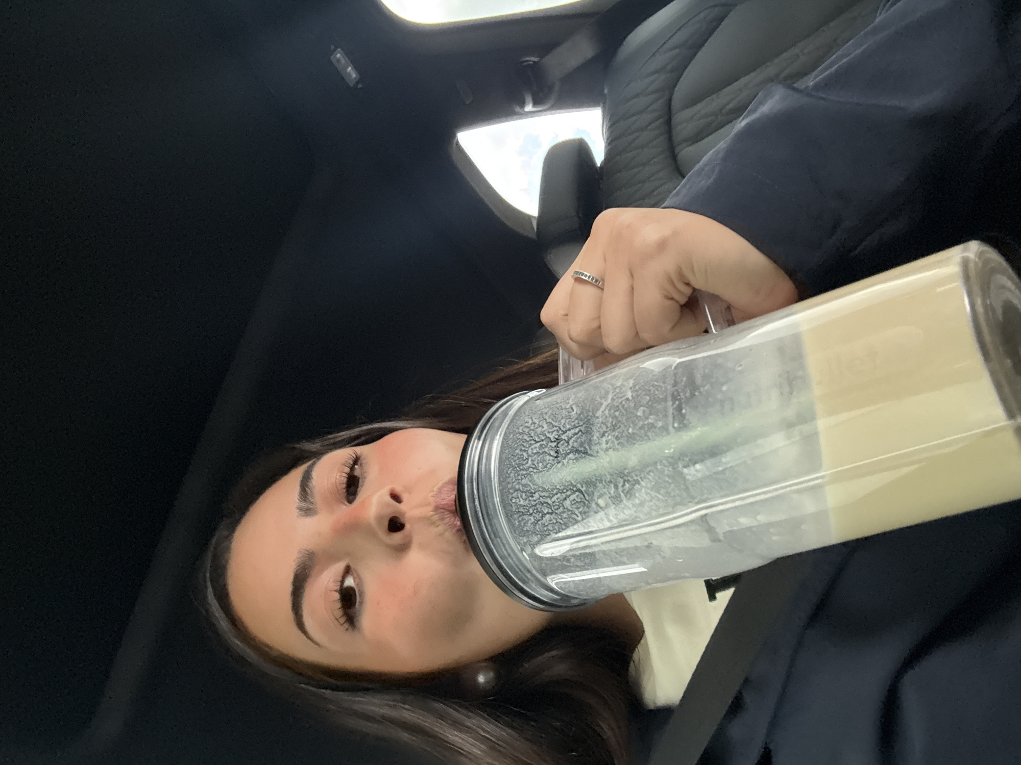 A woman is drinking from a clear plastic bottle containing a light-colored beverage, presumably a smoothie. She is likely in a car, indicated by the black interior with a sunroof above and a car seat to her side. The woman is light-skinned, with brown hair, wearing a dark-colored garment. She has a ring on her left hand. The context suggests the location might be in a vehicle, but there is insufficient information to definitively pinpoint the city of Ixtapaluca, Mexico. No additional details about the environment or additional actions can be determined from the image.