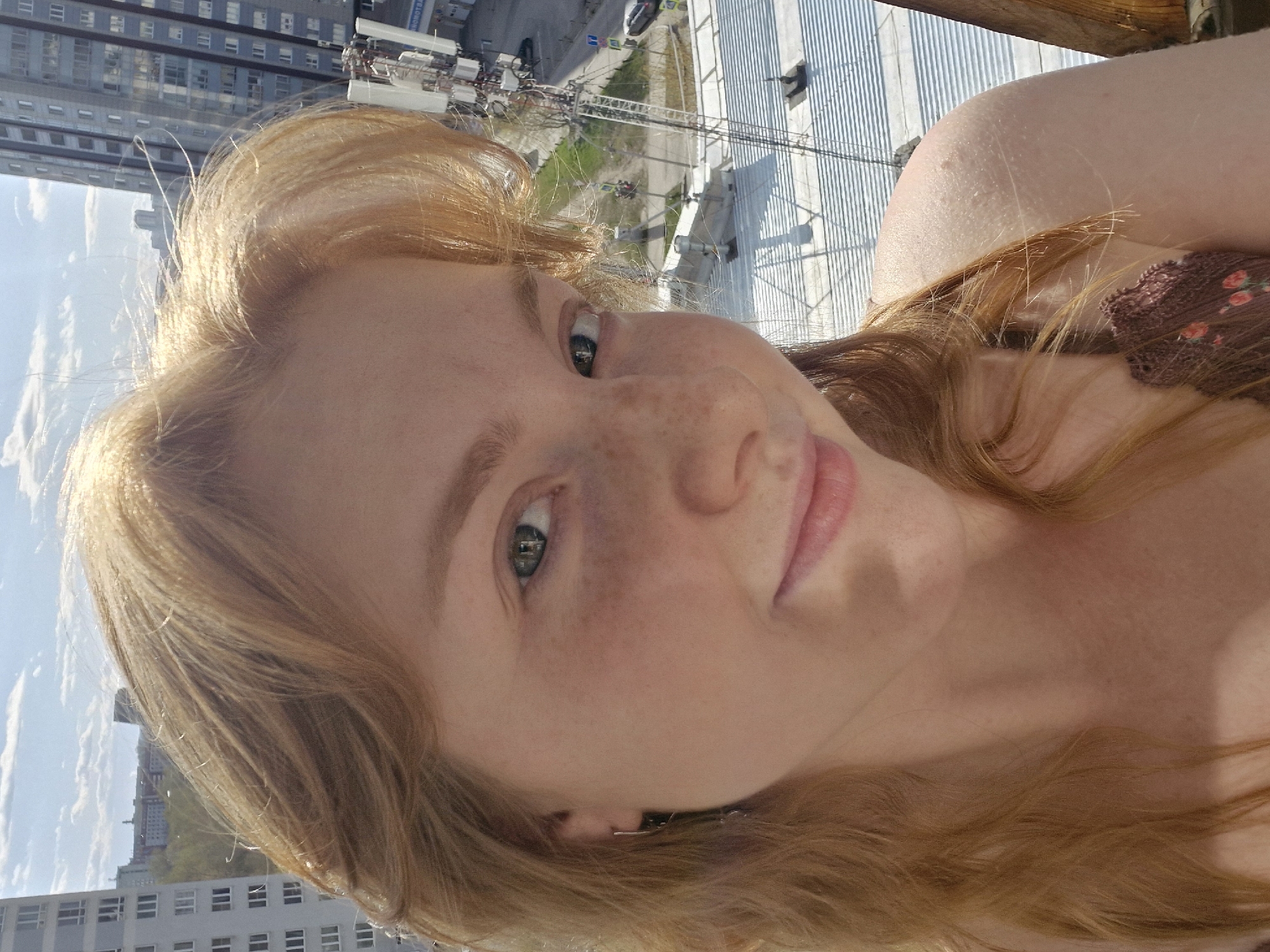 A close-up, rotated selfie features a young woman with fair, reddish-blonde hair and light skin, adorned with freckles across her nose and cheeks. Her striking green eyes gaze directly at the camera, and she presents a gentle, pleasant smile. A glimpse of a patterned top, possibly brown with floral details, is visible on her right shoulder.
The background reveals a panoramic urban landscape, suggesting the photo was taken from a high vantage point, likely a balcony or a tall building in Yekaterinburg, Russia. The perspective is tilted, showing the city at an angle. Numerous multi-story buildings with visible windows dominate the distant horizon, signifying a dense metropolitan environment. Below, there's a complex of urban structures, including what appear to be communication towers or industrial equipment, alongside a large, light-colored paved or corrugated surface that might be a rooftop or a large yard. A road with sparse vehicle traffic and some green areas snake through the urban infrastructure.
The image is captured in bright daylight, indicative of midday or afternoon, under a clear sky with a few scattered clouds. The strong sunlight highlights the woman's hair and face while casting distinct shadows across the background architecture. No discernible text is clearly readable within the frame. The scene conveys a casual outdoor portrait set against an active city backdrop.