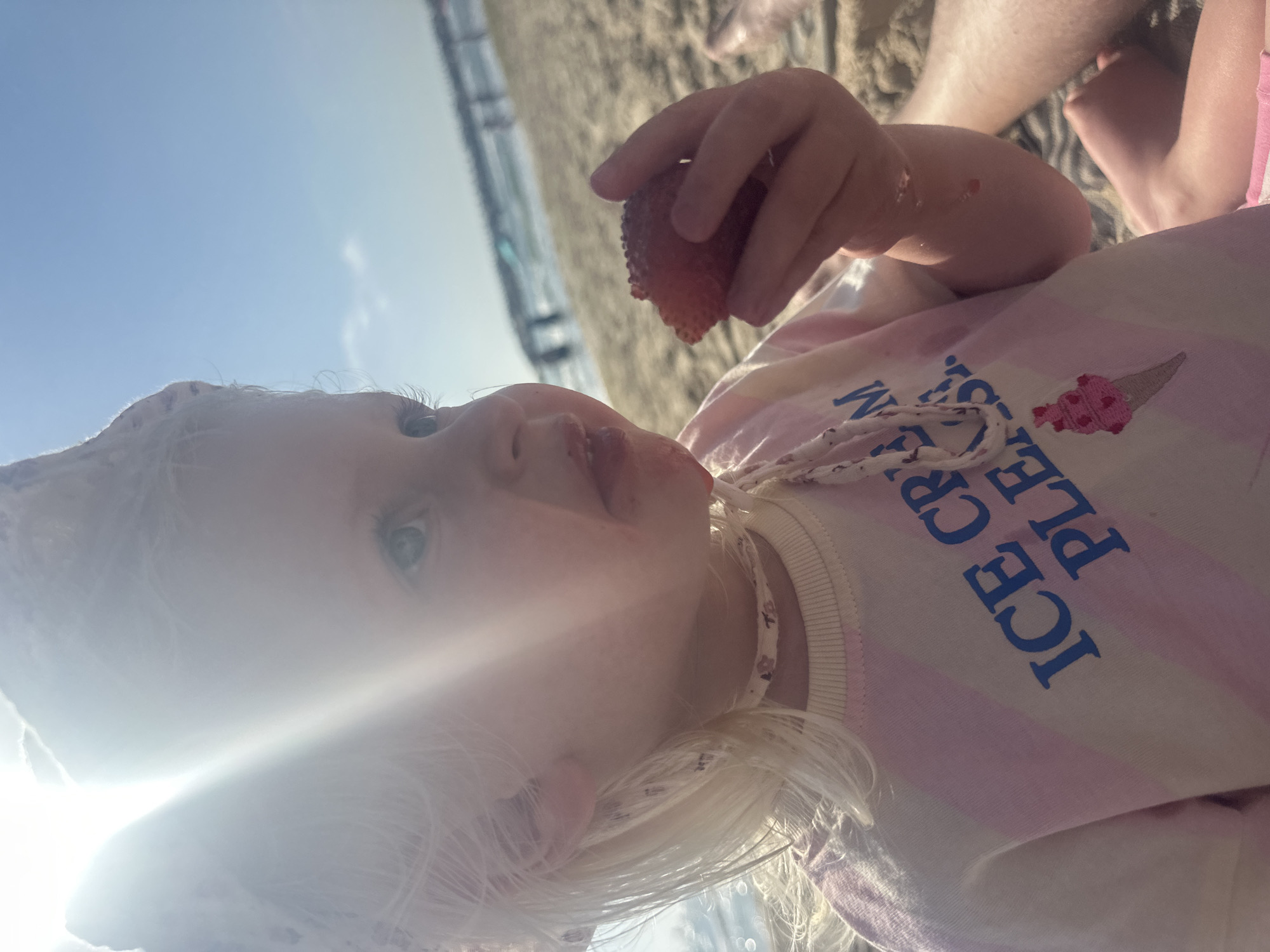 A young child with blonde hair and blue eyes is eating a strawberry on a beach. The child wears a light pink and white striped shirt with the words "ICE CREAM PLEASE" and a picture of an ice cream cone. They are holding a strawberry in their hand. Bare feet and legs are visible in the background. The photo appears to be taken on a sunny day at the beach, likely in Tel Aviv, Israel, as there is a building in the background.