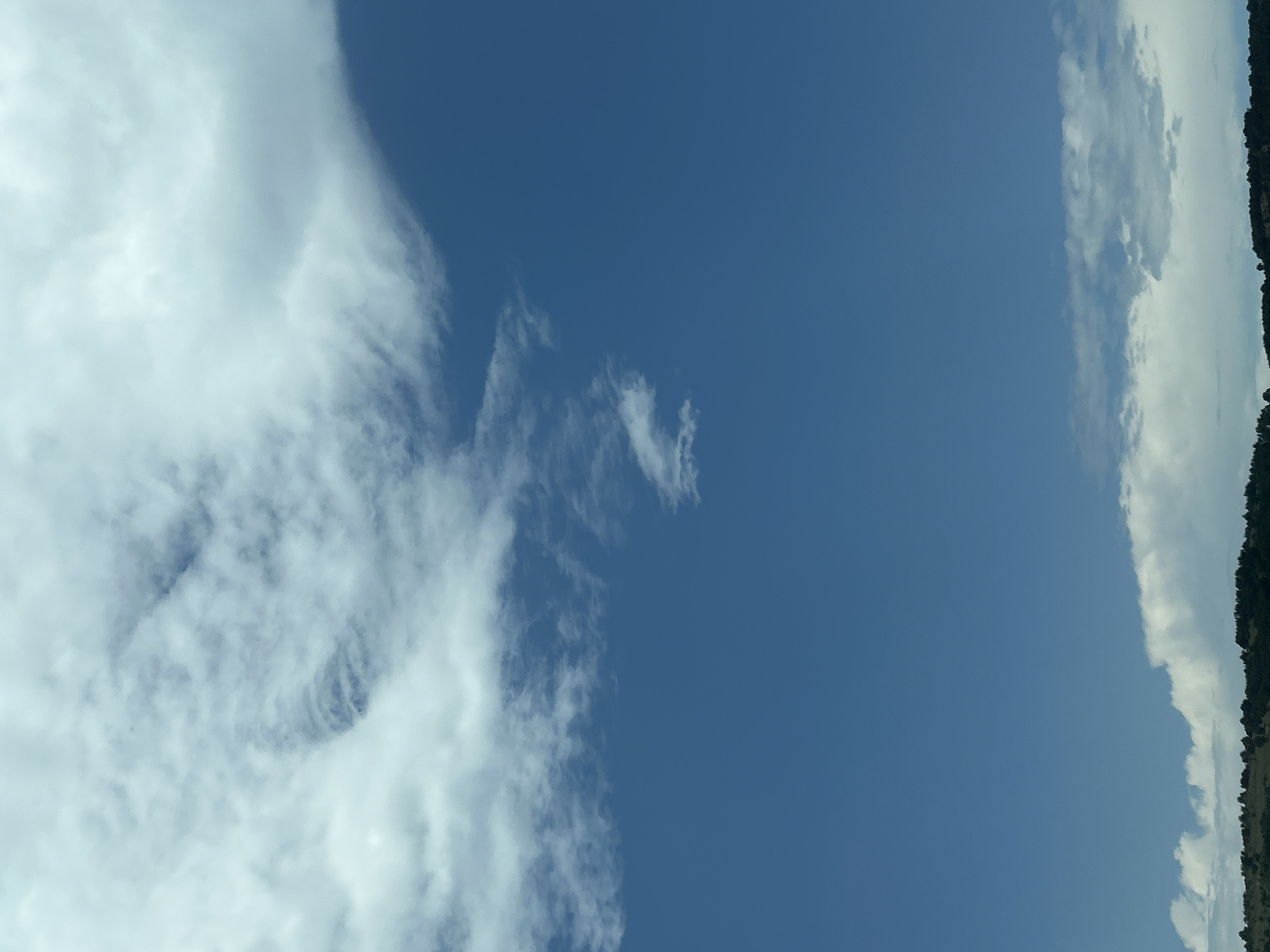 The image displays an expansive daytime sky over the distant landscape of Gorno Varshilo, Bulgaria. The upper third of the frame is dominated by a mass of white and light grey clouds, with a notable textured, swirling formation present in the upper-middle-left area. A sharp, diagonal edge of a cloud bank cuts across the upper-right quadrant, transitioning to a clear, deep blue sky that occupies the majority of the central and mid-right portions of the image. Along the lower horizon, a band of white and grey cumulus clouds is visible, resting just above a distant, dark green landscape characterized by undulating hills covered in dense foliage. No people, animals, man-made structures, or specific objects are discernible within the frame. The lighting indicates a bright, sunny day.
