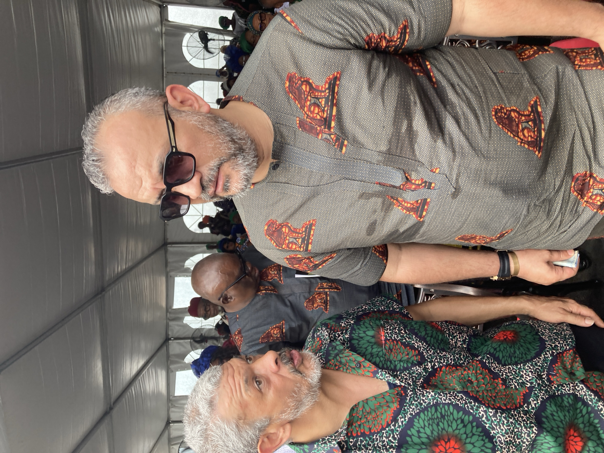 The image appears to be taken in a tent-like structure in Nnenu, Nigeria. At least three men are visible, likely part of a larger gathering of people. The man closest to the camera, is wearing a green shirt. The other man is wearing a gray shirt and sunglasses.