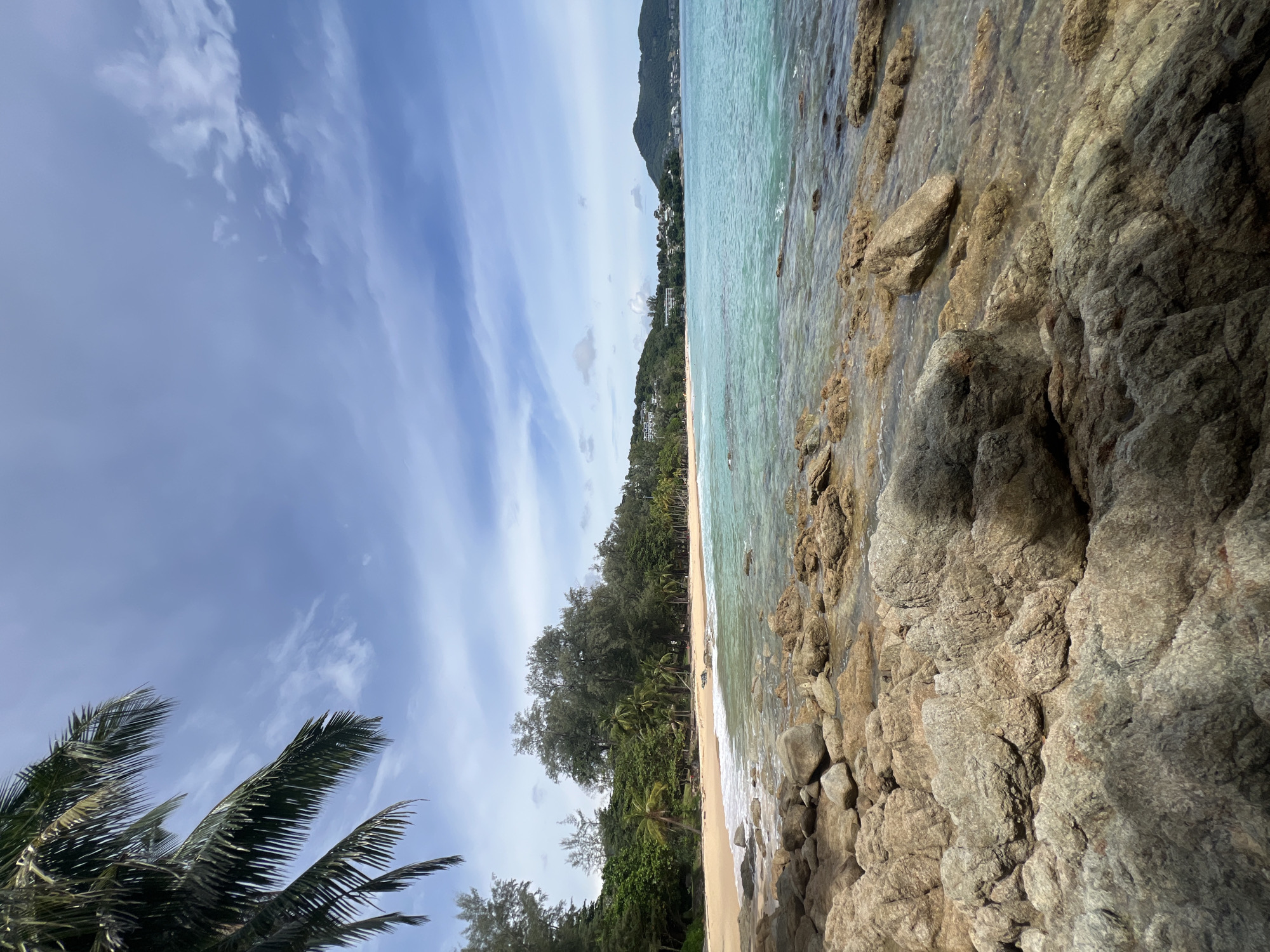 A wide-angle, eye-level shot captures a tropical beach scene in Choeng Thale, Thailand. In the foreground, large, weathered, tan-colored rocks are scattered across the shoreline and partially submerged in clear, shallow water. The water is a vibrant turquoise and exhibits gentle waves lapping at the shore. Beyond the rocky coast, a strip of golden sand beach is visible, backed by a dense line of green trees and palm trees, some with fronds reaching into the upper left portion of the frame. In the distance, along the coastline, there are hills covered in lush vegetation and dotted with buildings. The sky above is a clear blue with scattered white clouds. No people are present in the image.