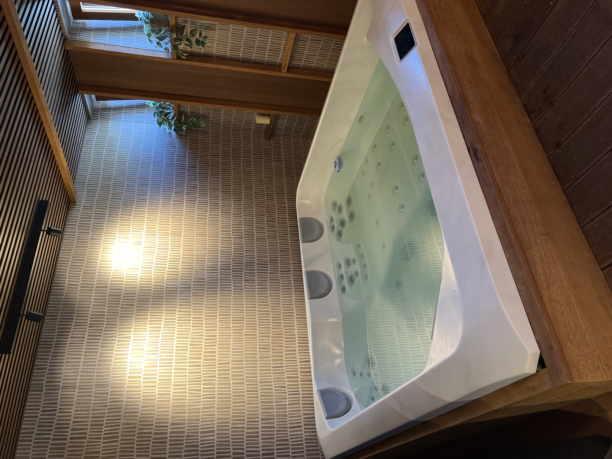 The image shows a well-lit, modern spa area, likely within a hotel or private residence in Mengusovce, Slovakia. Dominating the foreground is a large, white, built-in jacuzzi filled with clear, slightly rippling water. The jacuzzi has multiple jets visible beneath the surface and a digital control panel on its right side. The tub is surrounded by a wooden structure, and the floor is made of dark wooden planks. The background features a textured, neutral-toned wallpaper with a repeating vertical line pattern. Above the wallpaper, a slatted wooden ceiling with track lighting is visible. To the right, wooden panels create a partition, revealing glimpses of windows with greenery, possibly potted plants, on ledges. A small, unlit candle sits on a shelf within this wooden structure. The overall impression is one of relaxation and contemporary design.
