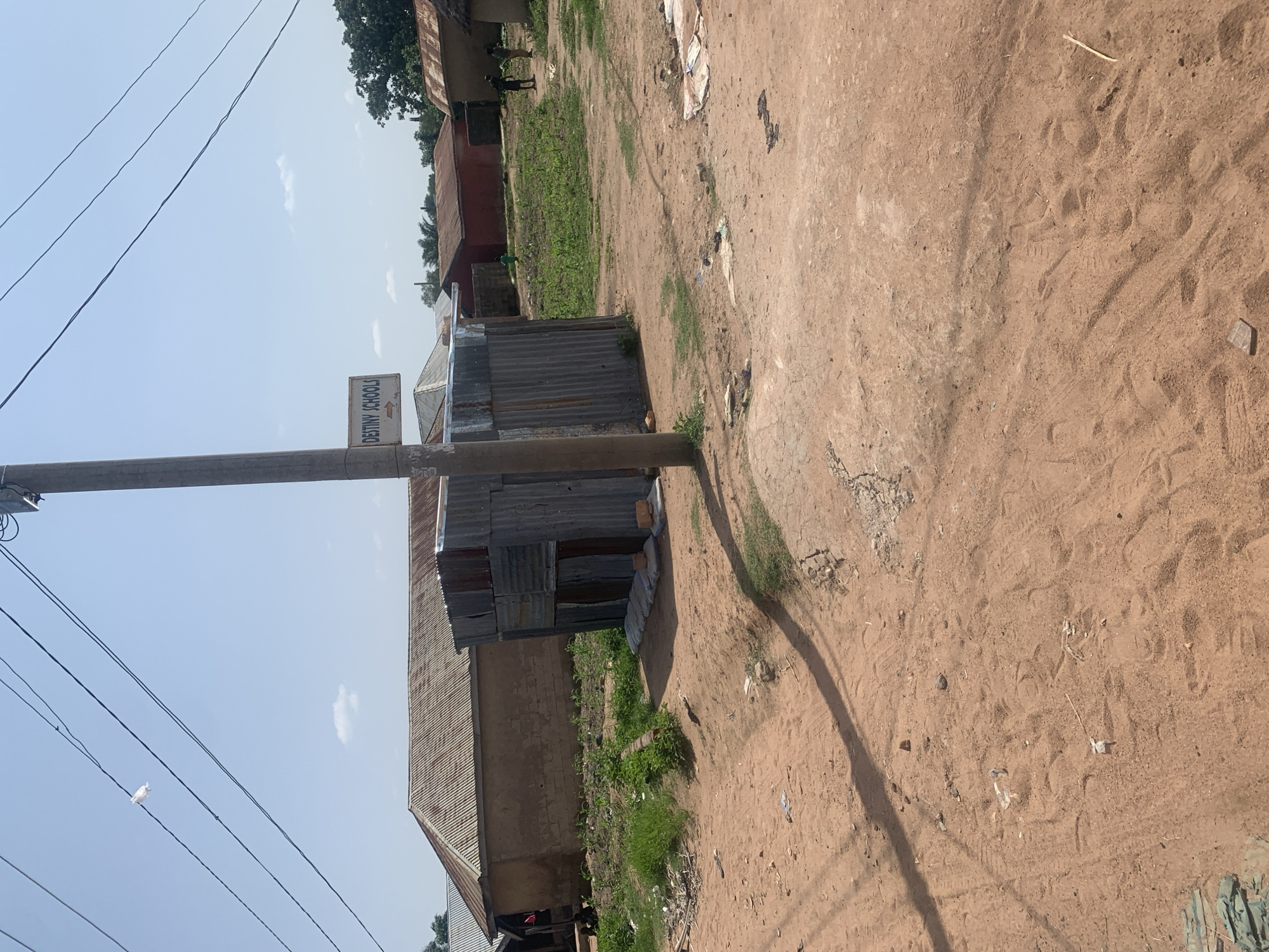 The image depicts an outdoor scene in Jalingo, Nigeria, with a focus on a dirt road and surrounding structures. The sky is clear with a few wispy clouds, suggesting it is daytime, likely morning or afternoon given the bright sunlight and defined shadows.
In the foreground, a dusty, sandy road occupies a significant portion of the frame. Tire tracks and footprints are visible in the dirt, indicating recent activity. To the left, a utility pole with several black wires stretches across the scene. A small white bird is perched on one of the wires.
Mounted on the utility pole is a sign with blue lettering that reads "DESTINY SCHOOLS" and an orange arrow pointing to the right. This sign suggests the presence of an educational institution nearby.
Further into the background, various structures are visible. There are buildings with corrugated metal roofs, some appearing to be makeshift or temporary. One building, possibly part of the "DESTINY SCHOOLS" complex, has walls made of mud bricks and a metal roof. Adjacent to this, another structure is constructed entirely from corrugated metal sheets. Red-colored buildings with tin roofs are also present, suggesting a mix of permanent and more informal construction in the area.
A small patch of green vegetation, possibly a garden or crops, is visible between some of the buildings. In the distance, a few figures can be seen, but they are too indistinct to identify clearly. The overall impression is of a rural or semi-urban setting in Nigeria, characterized by its unpaved roads, simple architecture, and everyday life. The scene appears peaceful and ordinary.