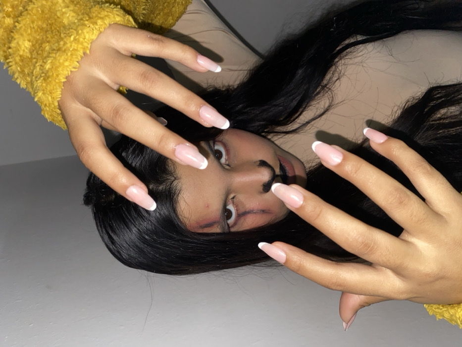A person with long black hair and long nails with white tips appears to be lying down. The individual wears makeup depicting a black nose and lines that extend from the nose across the cheeks. A yellow, fuzzy garment is partially visible in the upper left. The subject's eyes are wide and slightly startling. The background appears to be a plain, light-colored wall or surface.
