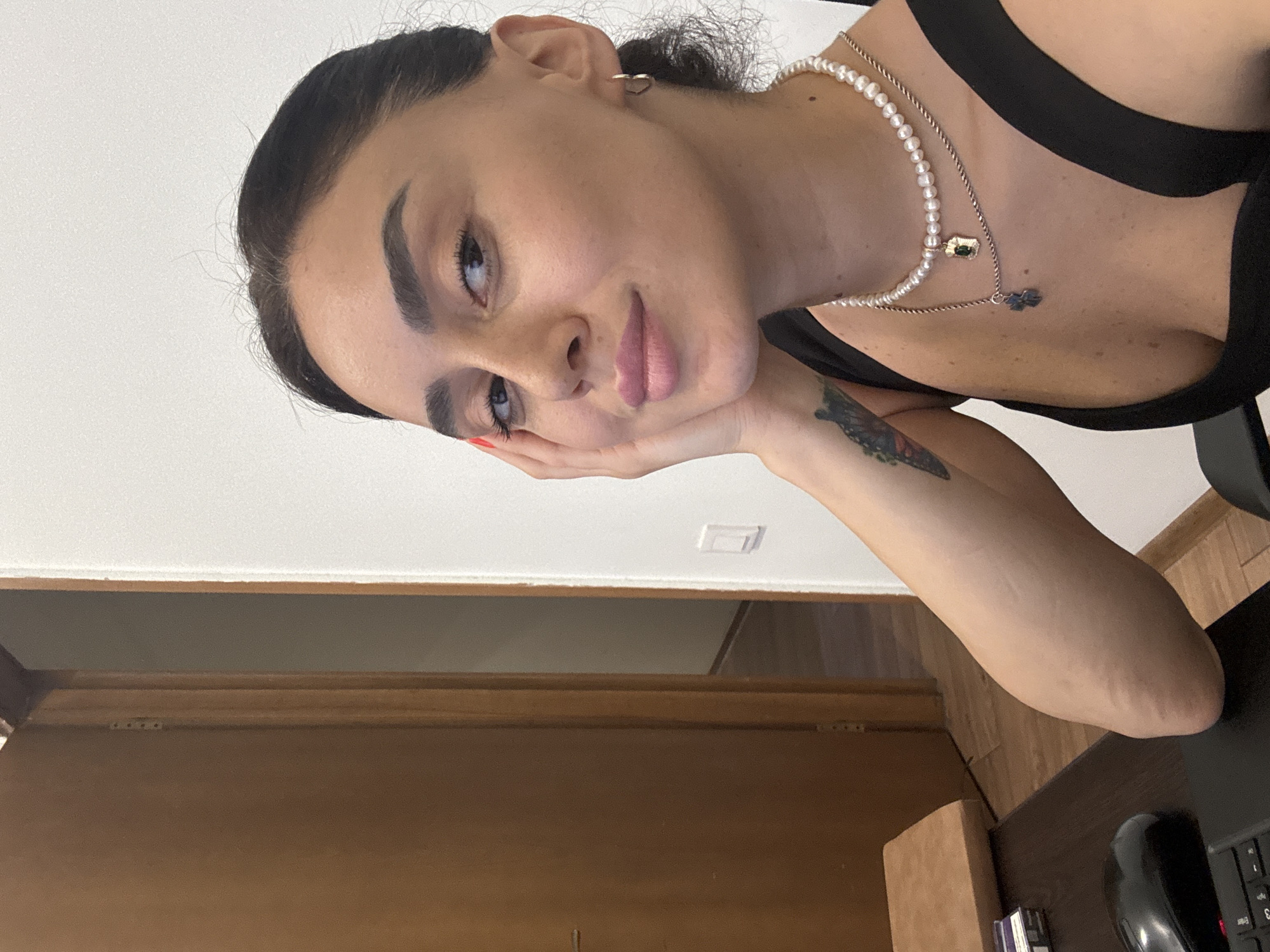 A woman is sitting at a desk in an indoor location. She has dark hair pulled back and is wearing a black top and a pearl necklace with two pendants. Her right arm is resting on her cheek. A door is visible in the background to the left, and a white wall with a light switch is to the right. The image was captured at a resolution of 1080x1350 pixels.
