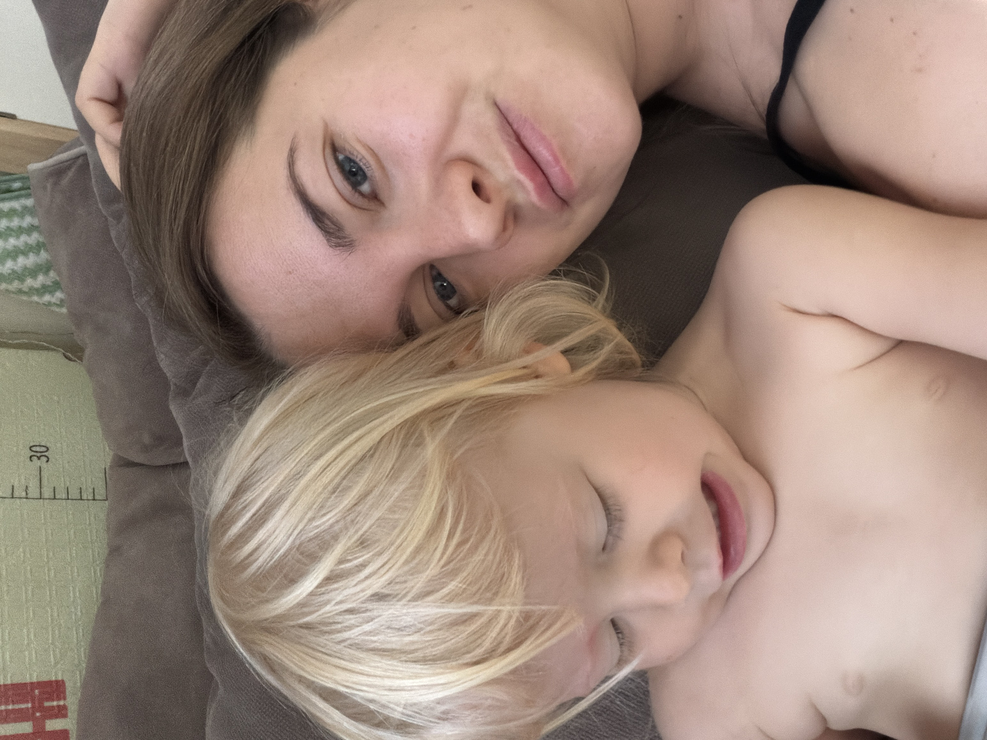 This media file captures an intimate indoor scene featuring an adult woman and a young child, likely in Sochi, Russia. Both individuals are lying down closely together on a soft, dark gray or brown surface, possibly a couch or bed.
The woman, with light brown hair and blue eyes, is positioned behind and slightly above the child. She looks calmly towards the camera with a gentle expression, wearing a dark-colored strap of clothing on her shoulder.
The young child, with bright blonde hair, is resting beside the woman. The child's eyes appear closed or nearly closed, and their mouth is slightly open, suggesting they are asleep or deeply relaxed.
In the background, to the left of the subjects, a light green mat with a patterned texture is visible. The mat displays measurement markings, including the number "30" and subsequent lines. Below this, partially visible red text, possibly "EH" or "IH," can be seen.
The soft, natural lighting suggests it is daytime. The overall mood of the image is peaceful and tender, capturing a quiet moment of rest or bonding between the adult and child.