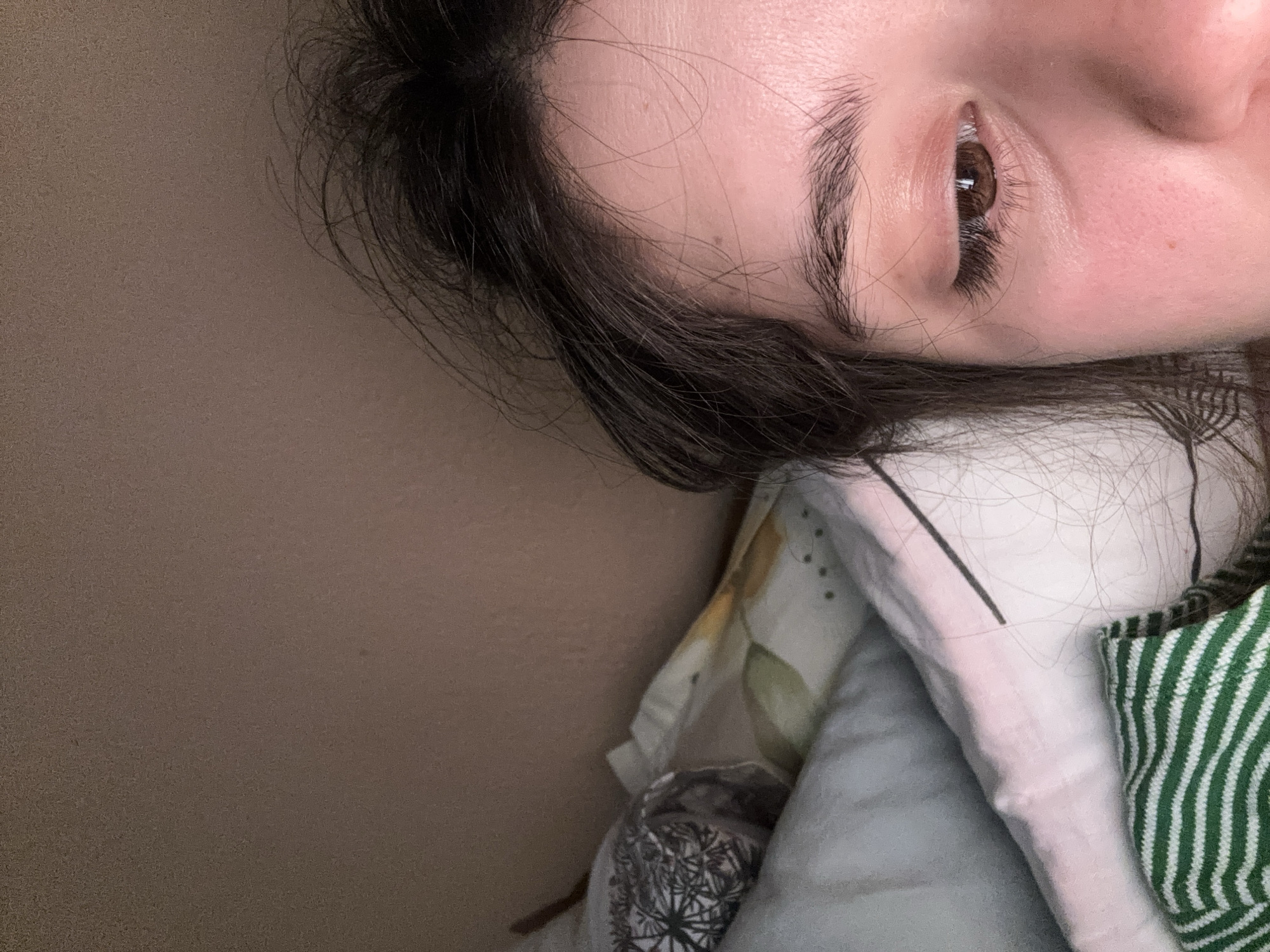 The image shows a partial view of a person lying down on what appears to be a bed or couch. The person's right eye, eyebrow, and forehead are visible. The eye is brown with visible eyelashes. Dark hair is present near the forehead. The person is lying on various pillows and blankets with different patterns, including a floral pattern, a black and white pattern, and a green and white striped pattern. The background is a solid brown wall. There is no indication of Garmen, Bulgaria, in this image.