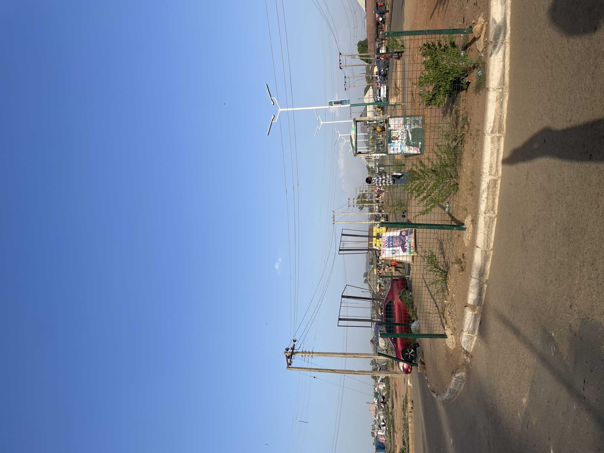 The image depicts an outdoor scene in Bukuru, Nigeria, featuring a street with a sidewalk separated by a green metal fence. Utility poles with power lines traverse the sky. Various posters are affixed to the fence. A red car is parked behind the fence. The street surface is asphalt with a concrete curb. A shadow is cast on the road. People are visible in the background. The sky is clear and blue.