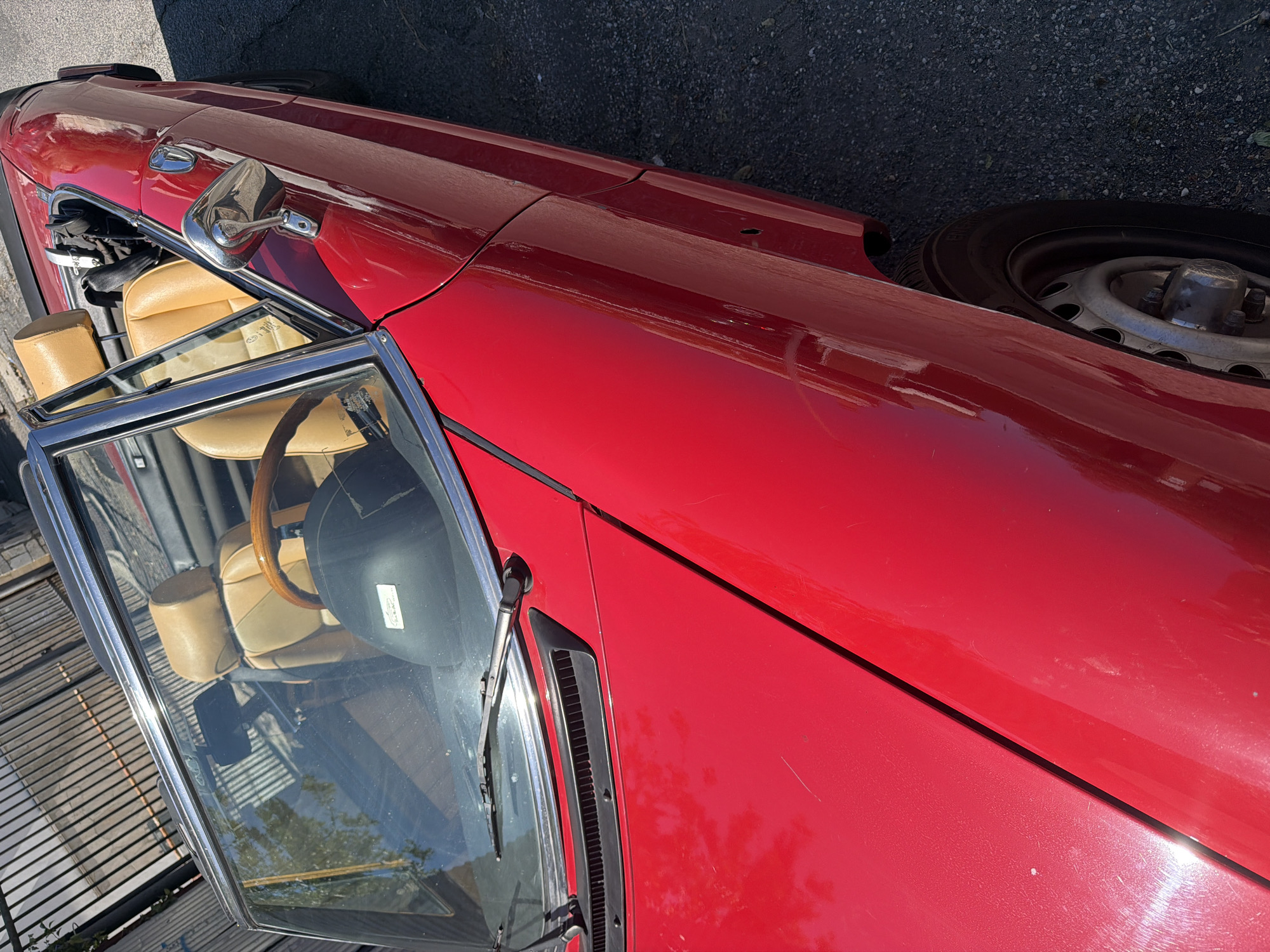 The image shows a close-up, high-angle view of a red convertible sports car parked on a paved surface, likely a street or parking lot in Rome, Italy. The car's interior features tan leather seats and a wooden steering wheel. A black helmet rests on the dashboard. The car's exterior is a vibrant red, with chrome accents on the side mirror. The background reveals a metal fence and some greenery, suggesting an outdoor urban setting. There are no people visible in the image. The lighting indicates it is daytime, with sunlight casting reflections on the car's polished surface. The asphalt ground is visible to the right of the car, appearing slightly worn.