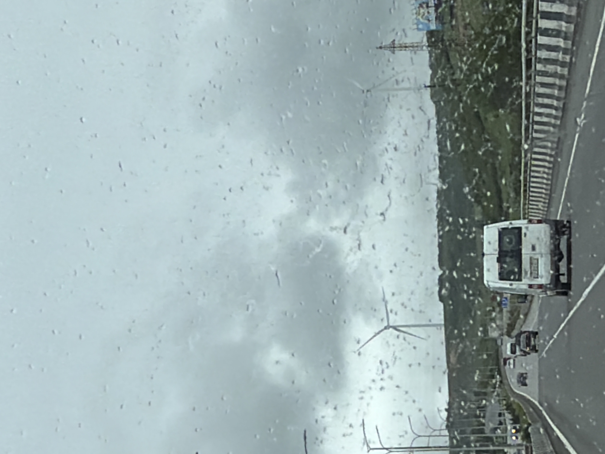 The image, taken through a rain-streaked window, depicts a view of a highway and landscape near Otarsheni, Georgia. Wind turbines are visible in the distance. There is a white van traveling on the right side of the road with other vehicles behind it. The side of the highway features a concrete barrier with black and white stripes. A green, hilly area borders the left side of the highway, with additional infrastructure, including streetlights, visible in the foreground. The sky is overcast.