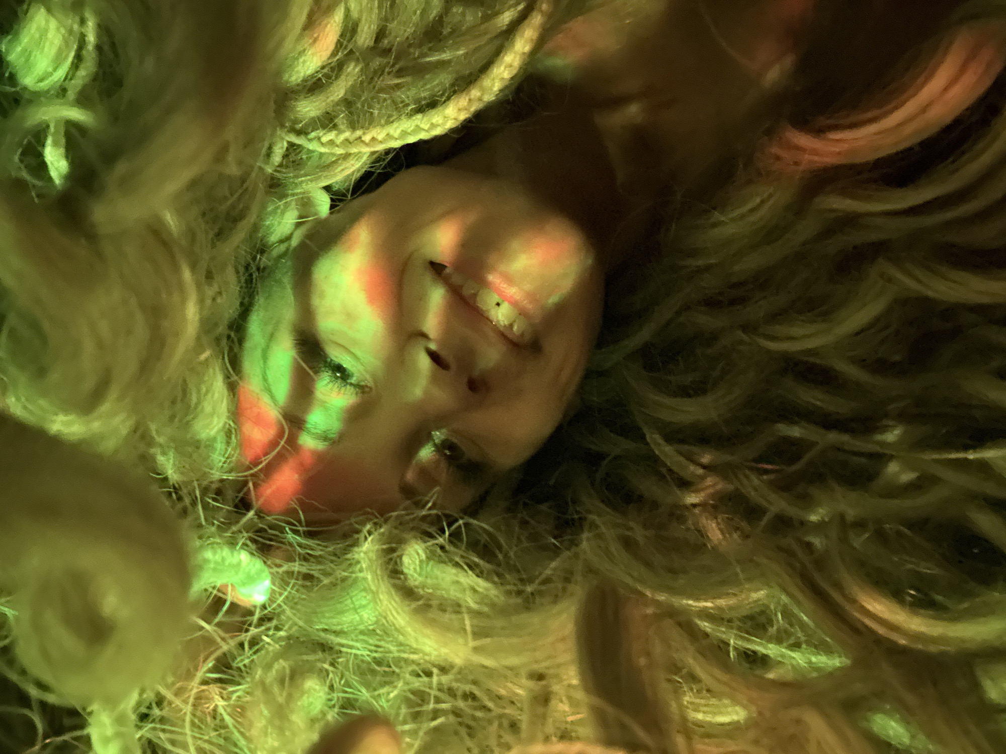 An image of a female face surrounded by light blonde hair, taken from an unusual angle and lit with green and red light. A braid is visible in the upper left corner of the image. The person is smiling slightly. The location in the city of Rusanovo, Russia, is not discernable in the image.